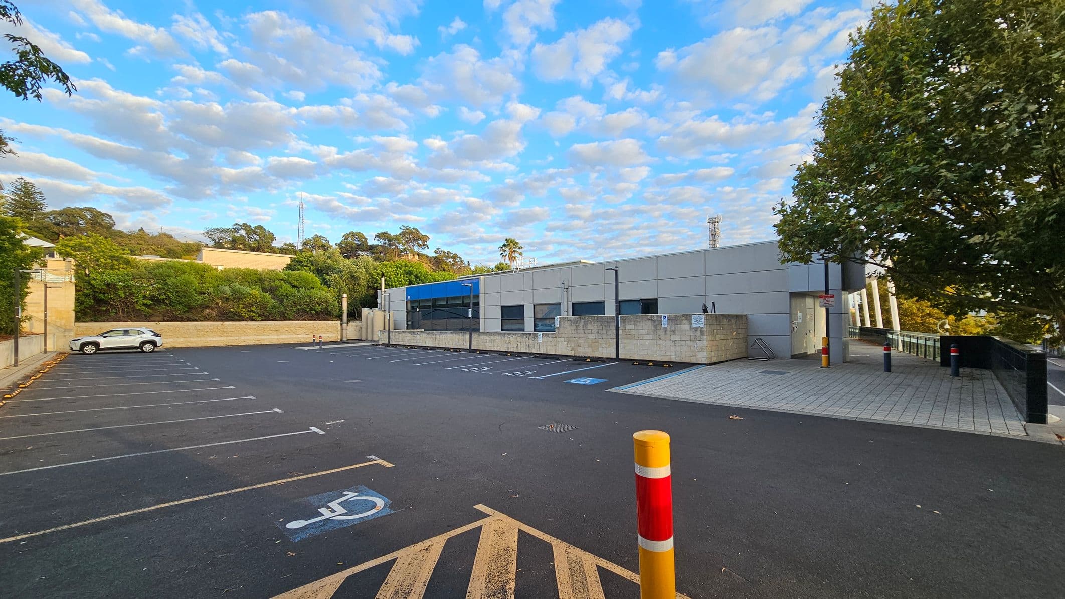 A car park with marked spaces, including a disabled parking spot in the foreground. On the left, a white car is parked near a stone wall with greenery and trees behind it. In the centre, a modern building with grey panels and blue accents is visible. On the right, a large tree provides shade over a tiled walkway leading to the building's entrance. A red and yellow bollard stands in the foreground, and several more are near the entrance.