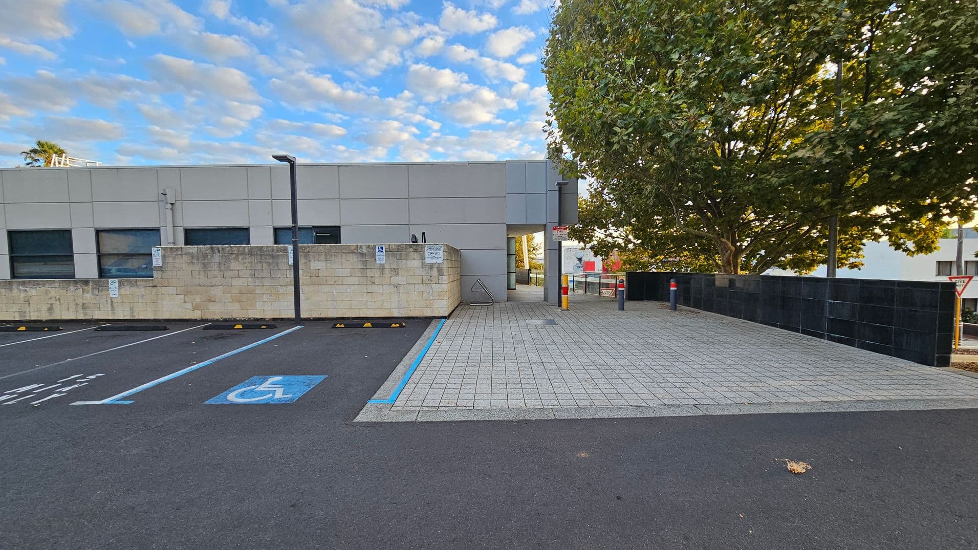 A paved area leads to a building entrance in the centre, with a large tree on the right providing shade. On the left, there are parking spaces marked with blue lines, including a designated disabled parking spot. A low wall separates the parking area from the building. The building has a modern design with large windows. A "Give Way" sign is visible near the tree. The ground is a mix of asphalt and paving stones.