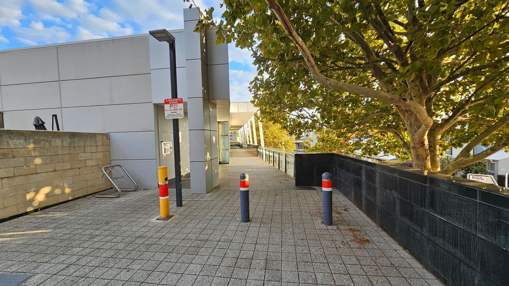 A paved walkway with grey tiles leads towards a modern building on the left, featuring a light grey facade. A signpost with a "Loading Zone" sign is visible near the building. On the right, a large tree with dense green foliage provides shade. Two bollards with red and white markings are positioned on the path. A black tiled wall runs parallel to the walkway on the right, and a metal bike rack is attached to a low brick wall on the left.
