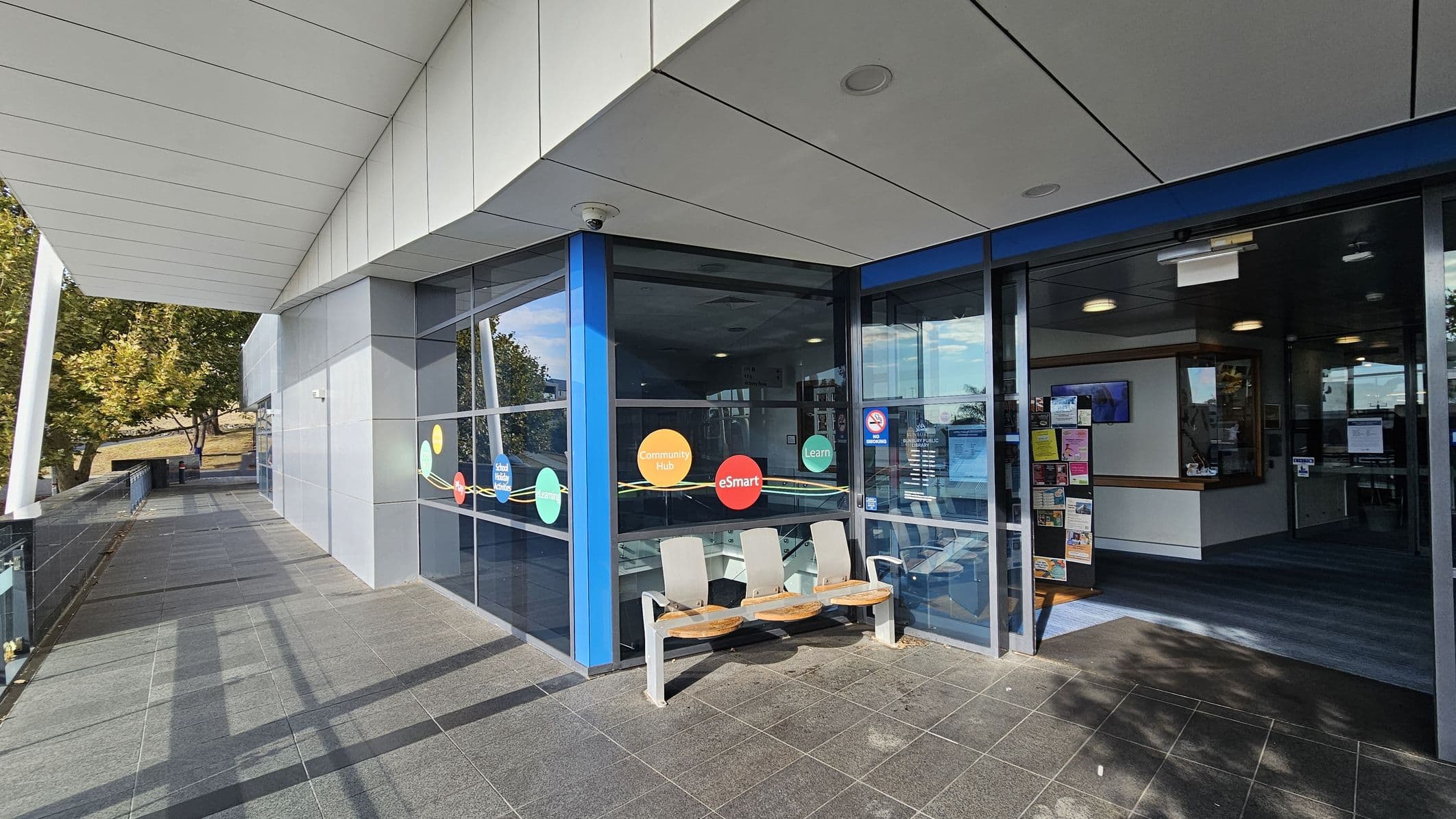A modern building entrance with large glass windows on the right, displaying colourful circular decals with text. Three white and wooden seats are positioned outside. The entrance door is open, revealing a reception area with posters and notices. The left side features a tiled walkway with trees in the background. The ceiling is white with recessed lighting, and the floor is grey tiled.