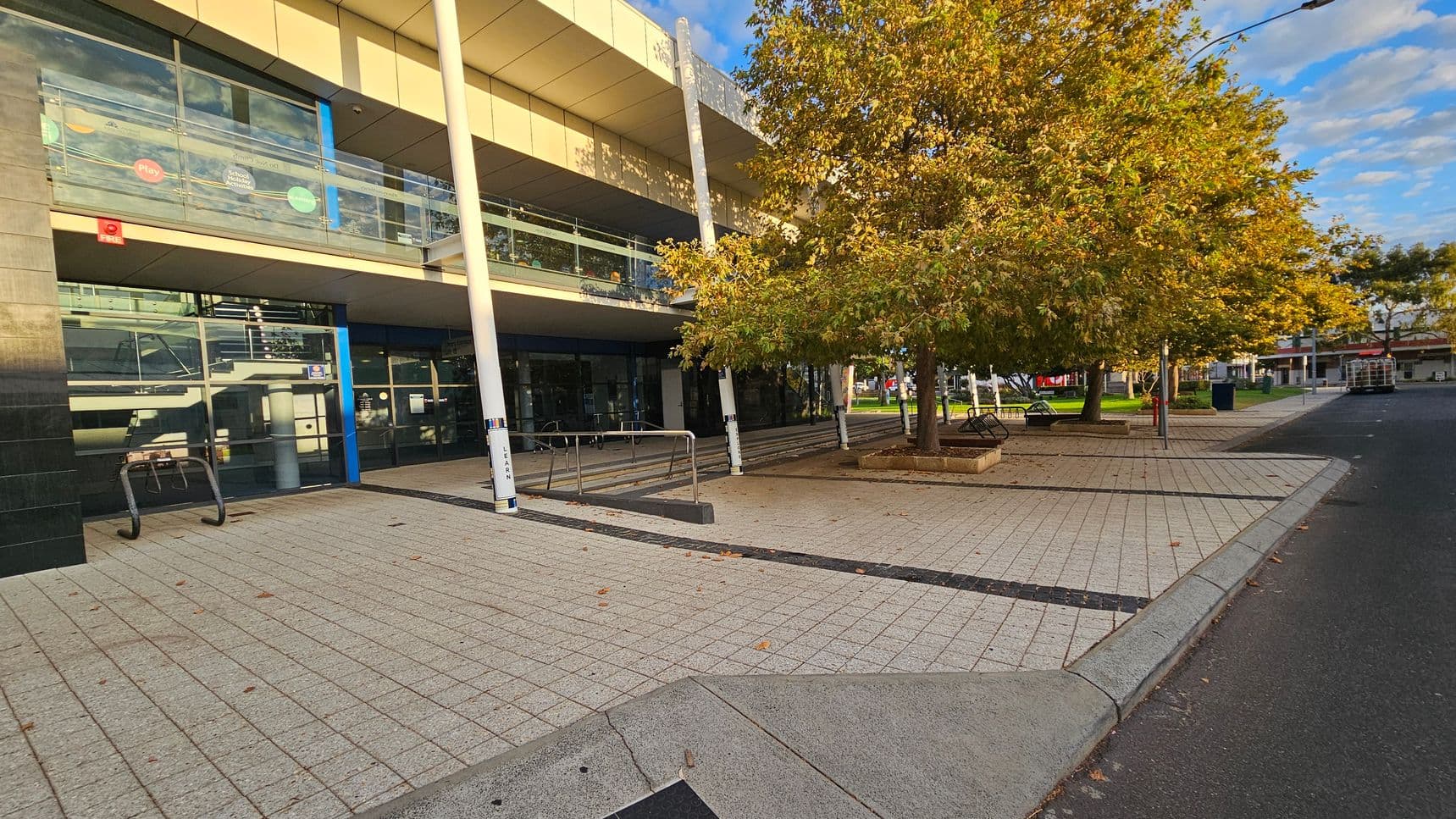 A modern building with large glass windows and a balcony is on the left. The entrance features a bike rack and a ramp with railings. On the right, there are several trees with autumn foliage, surrounded by paved walkways and benches. The ground is covered with light-coloured tiles, and a road runs alongside the pavement. The ceiling of the building is flat and beige, with white support columns.