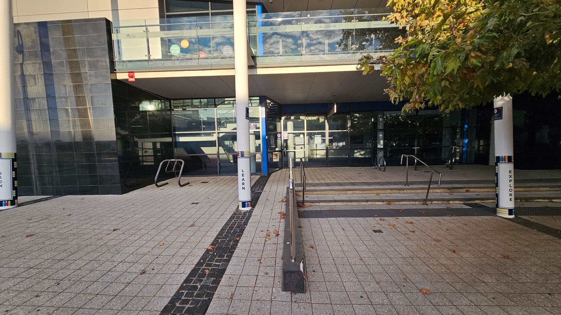 A modern building entrance with a tiled walkway leading up to it. On the left, there is a black tiled wall and a bicycle rack. In the centre, steps and a ramp with metal railings lead to glass doors. Two white pillars with the words "LEARN" and "EXPLORE" are visible. On the right, a tree with green and brown leaves provides some shade. The upper level features glass railings with various stickers. The ground is covered with grey and beige tiles, scattered with a few fallen leaves.