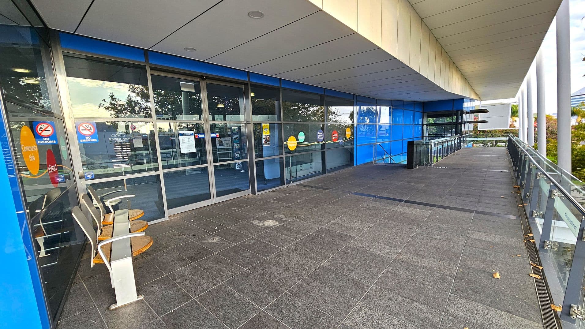 A modern building entrance with large glass doors and windows on the left, featuring colourful circular signs and "No Smoking" notices. Three chairs are positioned near the entrance. The floor is covered with dark grey tiles, and the ceiling is light-coloured with recessed lighting. On the right, a glass railing runs along the edge of a walkway, with trees visible in the background. The building exterior is clad in blue panels.