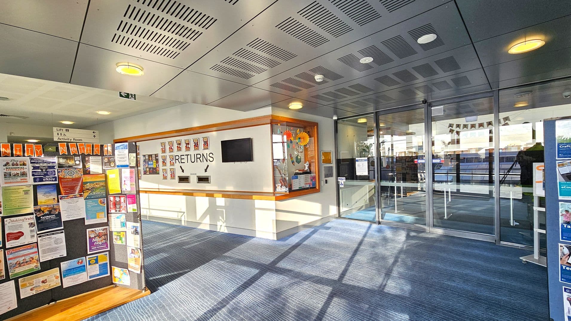 A library entrance area with a blue carpeted floor. On the left, a community notice board displays various colourful posters and flyers. In the centre, a wall marked "RETURNS" features two book return slots and a small screen above. To the right, a glass entrance door leads into the library, with a display case beside it containing decorative items. The ceiling is grey with recessed lighting and ventilation panels.