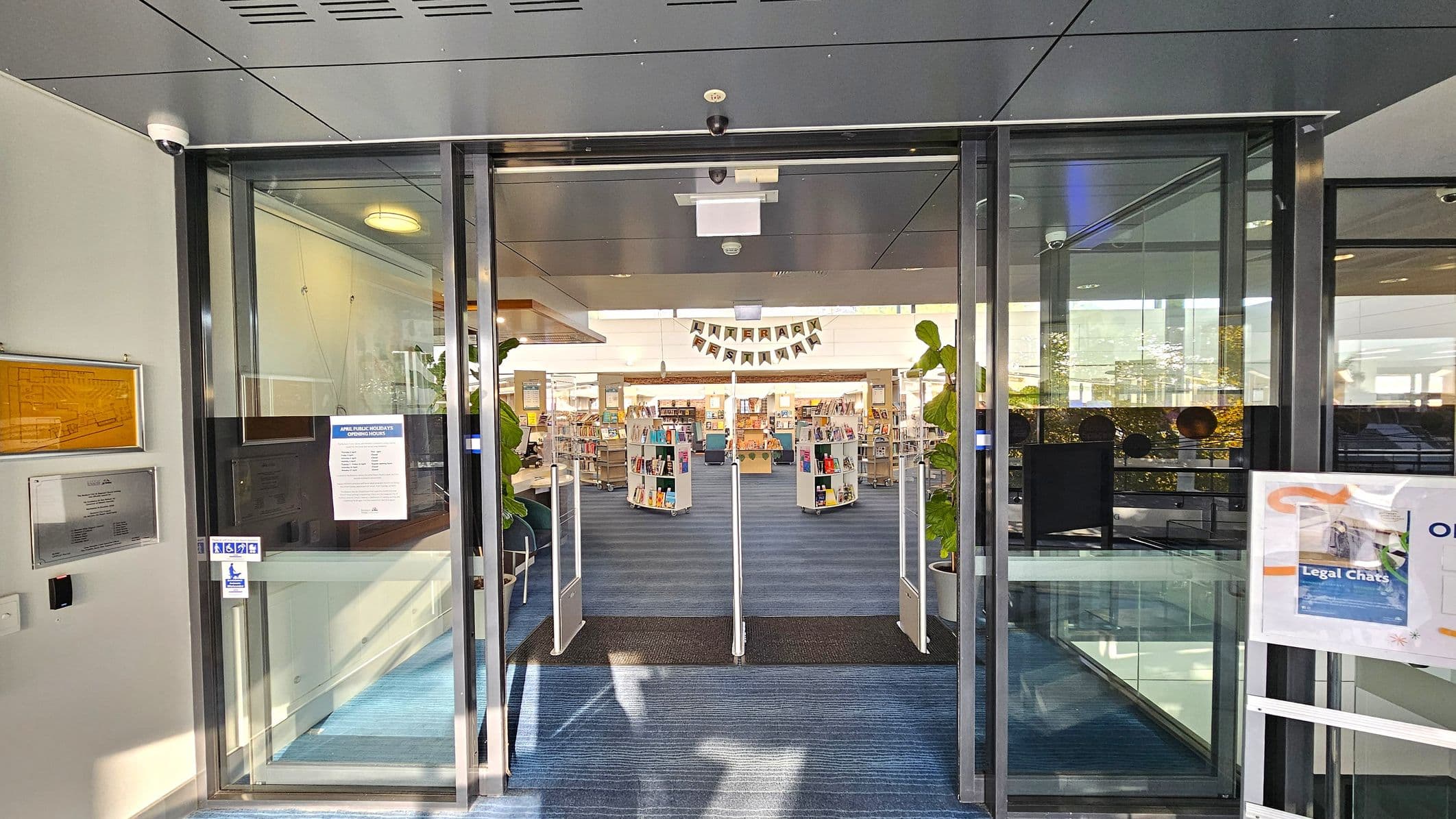 Glass entrance doors to a library with a blue carpeted floor leading inside. On the left, there are notices and a sign with opening hours. On the right, a poster is displayed on a stand. Inside, bookshelves are visible with books arranged on them. Large potted plants are placed near the entrance. The ceiling is grey with recessed lighting. A banner hangs in the background, and the interior is well-lit with natural light.