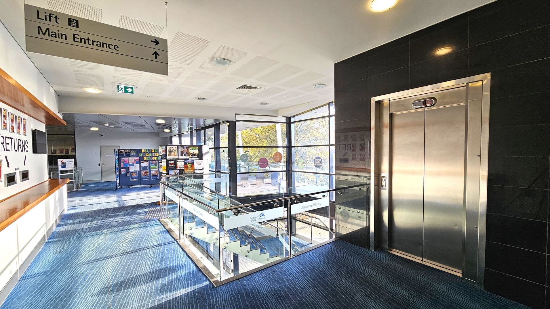 On the right, there's a shiny metal lift with a black tiled wall. In the middle, a glass-panelled staircase leads downwards, surrounded by large windows showing trees outside. On the left, a wall with a wooden ledge has a "RETURNS" slot and posters. Above, a sign points to the lift and main entrance. The floor is covered with blue carpet, and the ceiling is white with lights and vents.