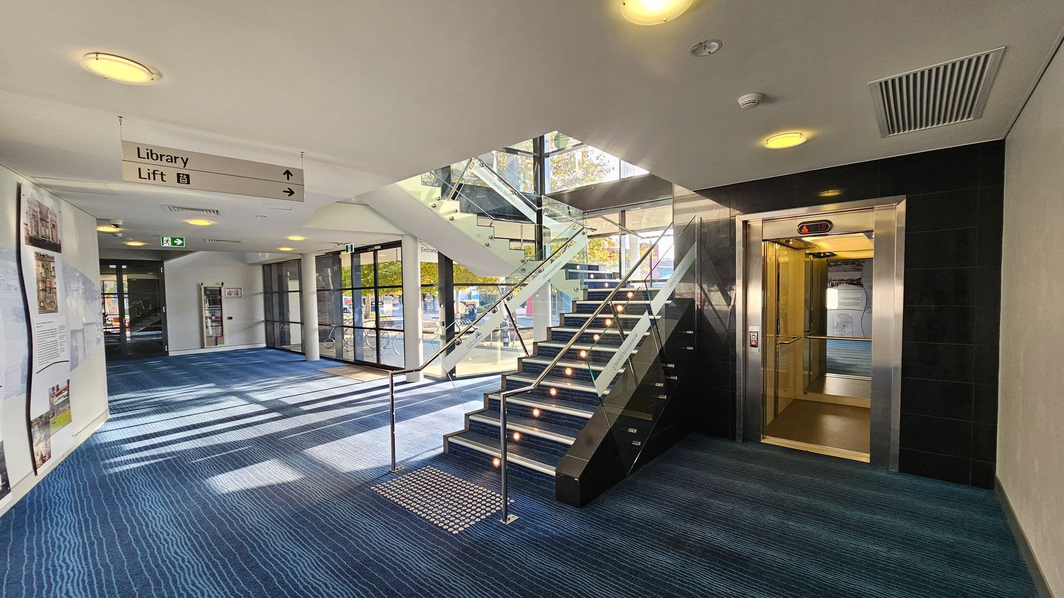A modern indoor space with a blue-patterned carpet. On the right, there's an open lift with metallic doors. In the centre, a staircase with glass railings leads upwards. To the left, large windows allow natural light to flood in, and there's a wall with informational displays. A ceiling sign indicates directions to the library and lift. The ceiling is white with recessed lighting and ventilation panels.