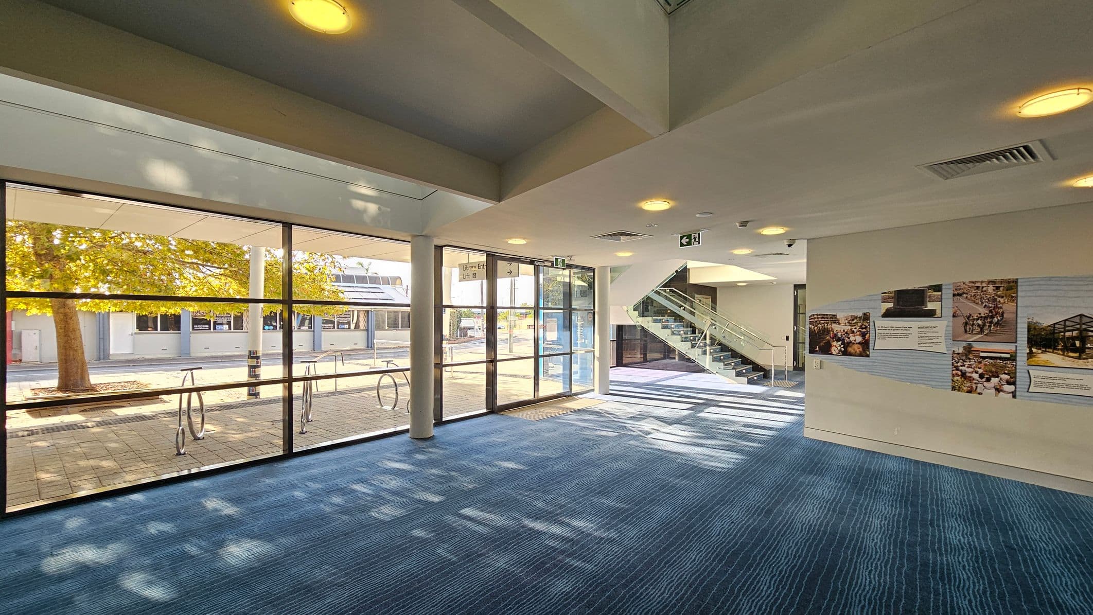 A spacious interior with large glass windows on the left, revealing an outdoor view with trees and a paved area. The floor is covered with blue carpet, and the ceiling has recessed lighting. On the right, a wall displays several posters with images and text. In the centre, a staircase with glass railings leads to an upper level. The area is well-lit with natural light streaming in through the windows.