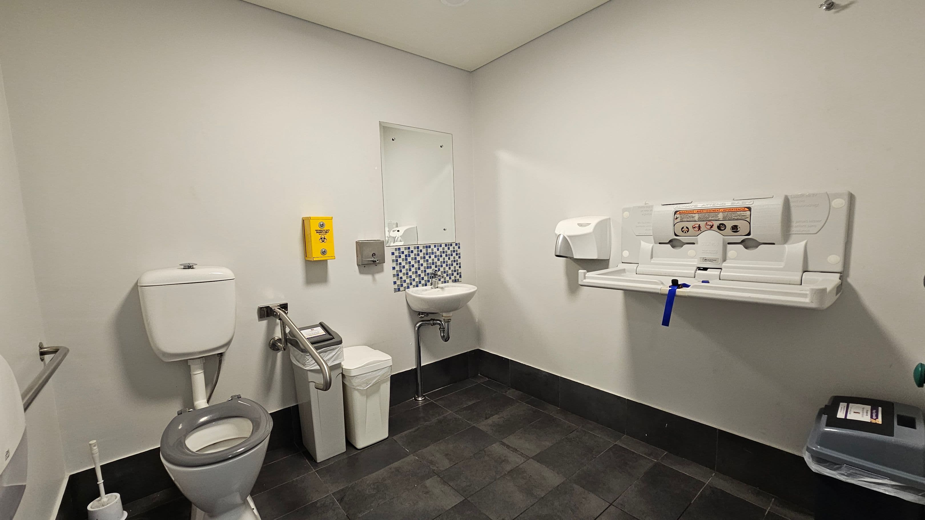 A public restroom with a grey tiled floor and light-coloured walls. On the left, there is a toilet with a grey seat, a toilet brush, and a grab rail. A yellow emergency button is mounted on the wall. In the centre, a small sink with a mirror above it is flanked by a soap dispenser. On the right, a baby changing station is mounted on the wall, with a paper towel dispenser nearby. A bin is positioned below the sink and another bin is on the far right. The ceiling is plain and light-coloured.