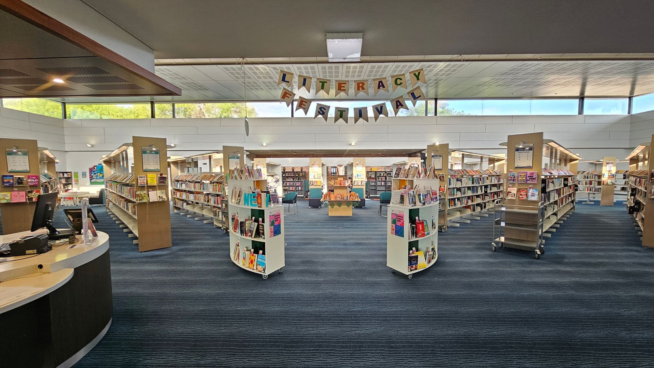 A library interior with a "Literacy Festival" banner hanging from the ceiling. In the foreground, there are curved bookshelves filled with books on either side. The middle ground features more bookshelves arranged in rows, leading to a central area with chairs and a table displaying books. The floor is carpeted in blue tones, and the ceiling has a grid pattern with fluorescent lights. Large windows near the ceiling allow natural light to enter.