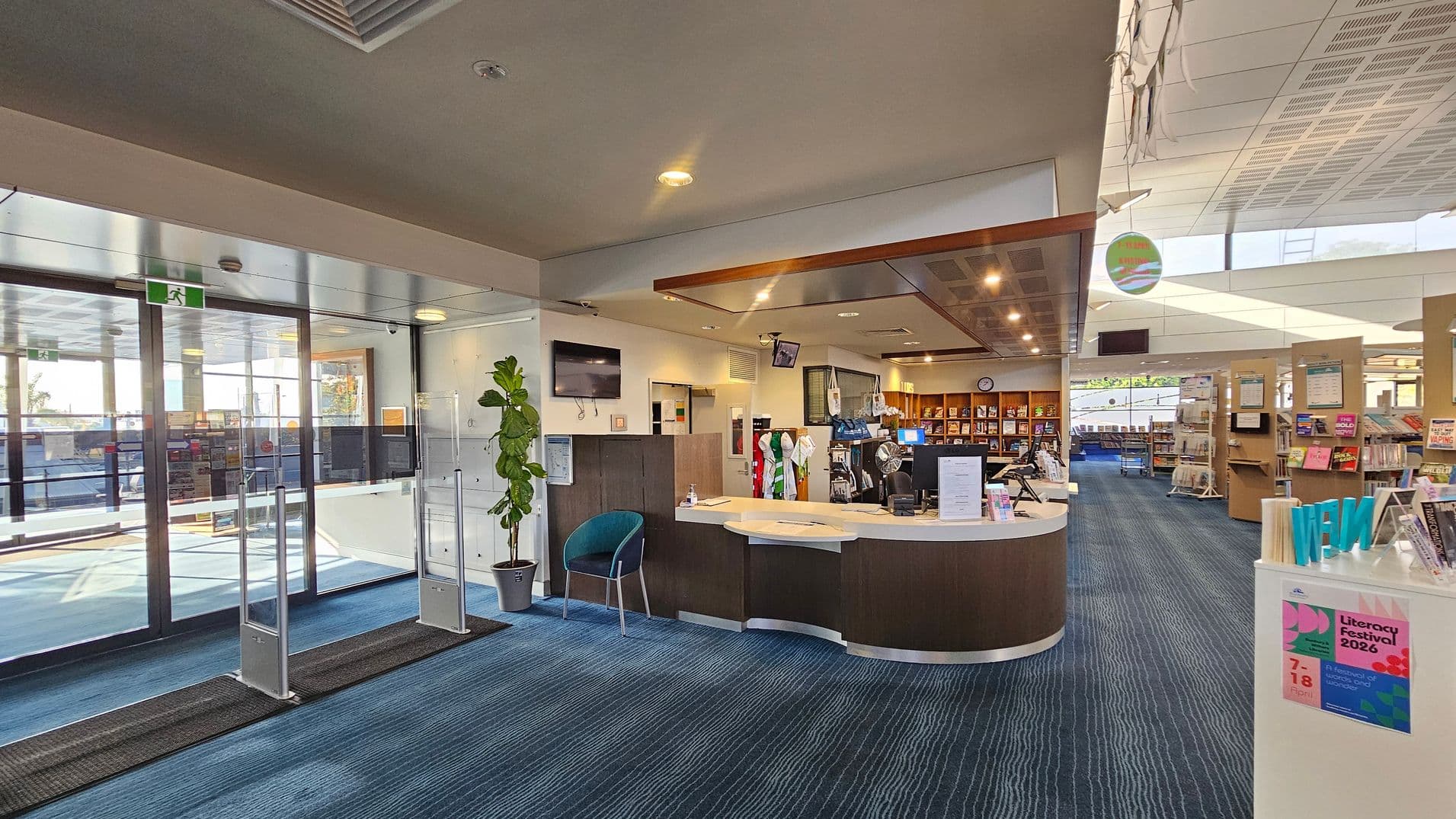 A library interior with a blue carpeted floor. On the left, there are glass entrance doors with a security sensor and a potted plant nearby. In the centre, a curved reception desk with computers and a chair is visible. Behind the desk, shelves filled with books and a wall-mounted TV can be seen. On the right, there are more bookshelves and a display stand with posters. The ceiling has recessed lighting and a modern design.