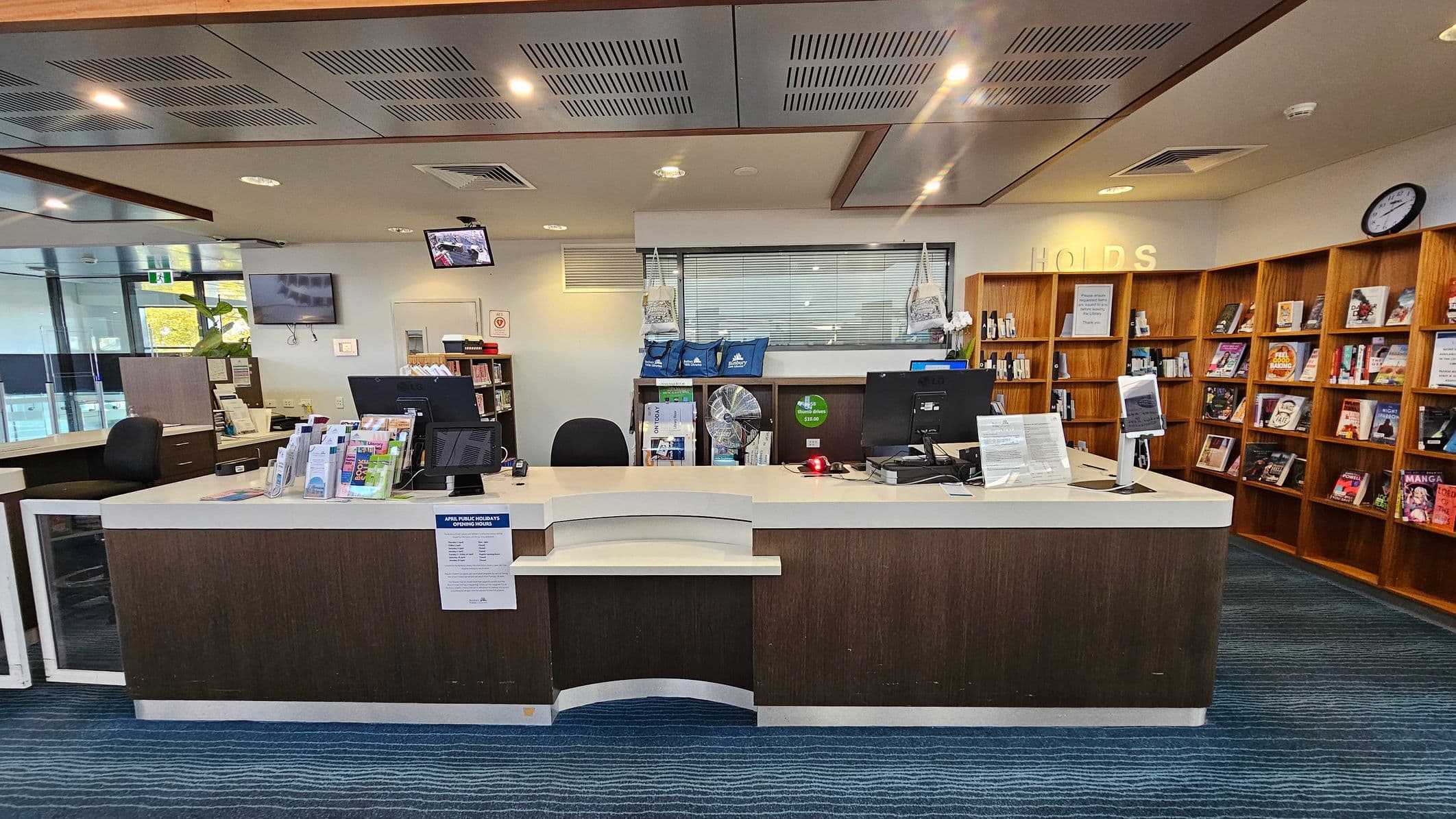 A library counter with a curved design is at the centre, featuring computers, brochures, and a sign with opening hours. Behind the counter, there are shelves with blue bags and a window with blinds. To the right, wooden bookshelves display various books. The ceiling has recessed lighting and ventilation panels, while the floor is carpeted in a blue pattern. A clock is visible on the wall to the right.