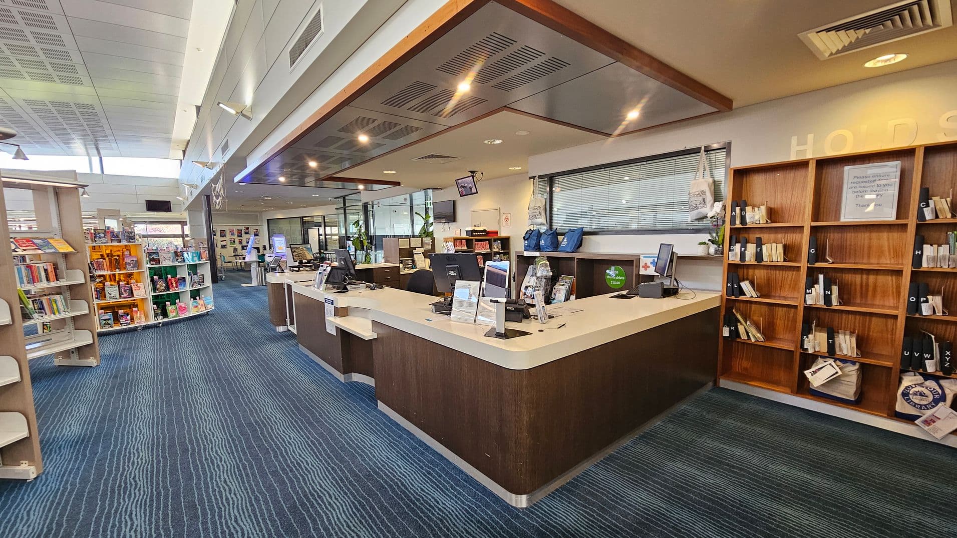 A library interior with a modern design. On the right, there is a wooden counter with computers and informational brochures. Behind the counter, shelves hold books and bags. On the left, there are bookshelves filled with colourful books. The floor is covered with a blue carpet featuring wavy patterns. The ceiling is white with recessed lighting and air vents. In the background, more bookshelves and a seating area are visible.
