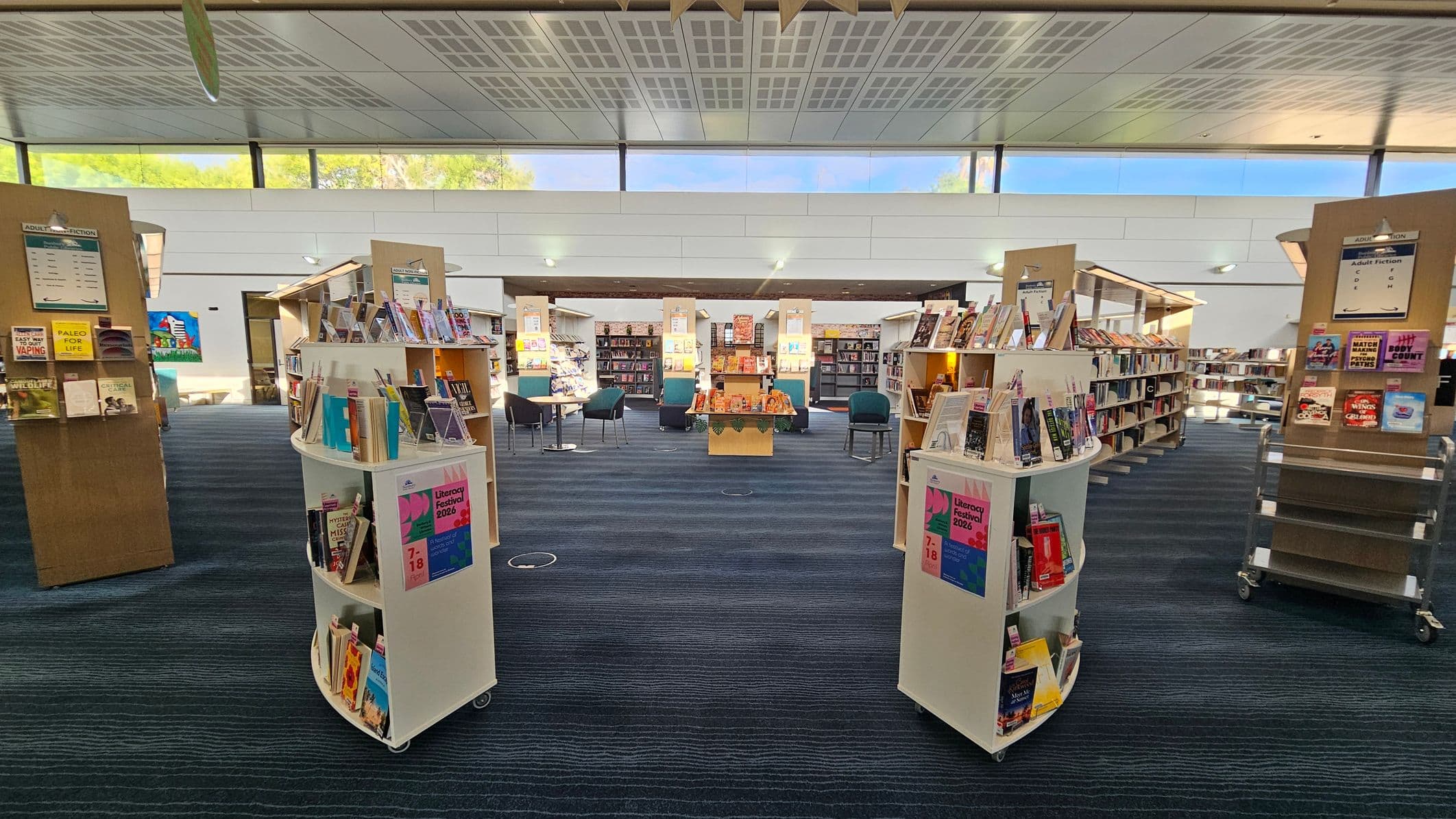 A library interior with a modern design. In the foreground, two white, curved bookshelves display various books and a poster for a Literacy Festival 2026. The middle area features more bookshelves and a central table with chairs around it. The background shows additional shelves filled with books. The ceiling is patterned with square panels, and large windows near the top allow natural light to illuminate the space. The floor is carpeted in dark blue with subtle patterns.
