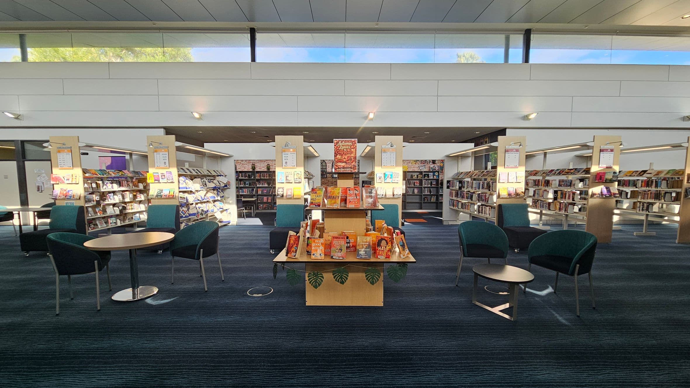 A library interior with a central display table featuring books with orange covers. Surrounding the table are several green chairs and round tables. In the background, there are bookshelves filled with books, and the walls are lined with more shelving units. The ceiling is high with large windows allowing natural light to illuminate the space. The floor is carpeted in a dark blue and green pattern.
