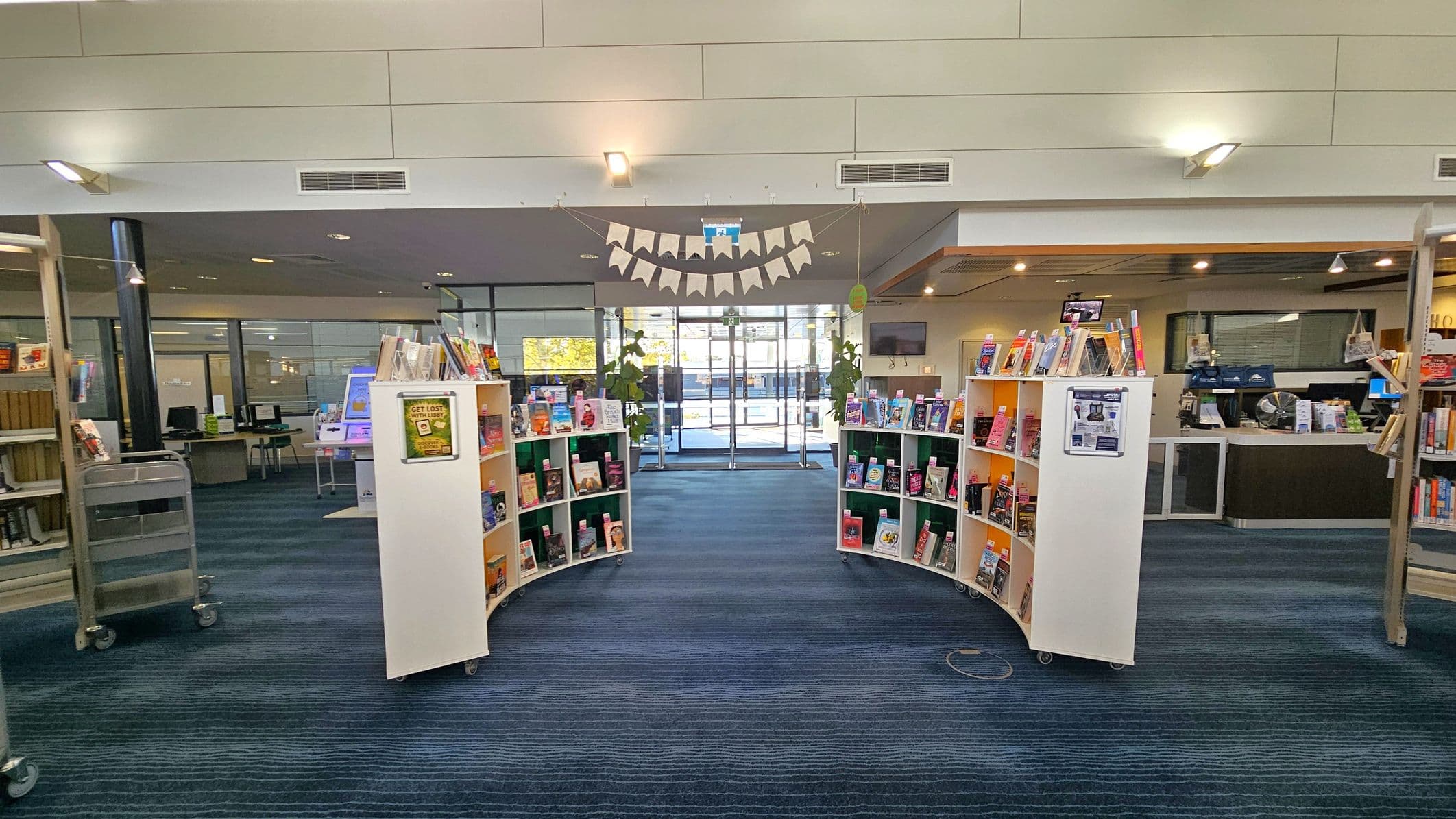 Curved white bookshelves filled with books are positioned in the centre, creating a pathway leading to glass doors in the background. Above, a string of triangular flags is suspended. The floor is covered with blue carpet, and the ceiling features recessed lighting. On the left, there are computer stations and a book trolley. To the right, a counter area with more books and a monitor is visible. Large windows and plants are near the entrance.
