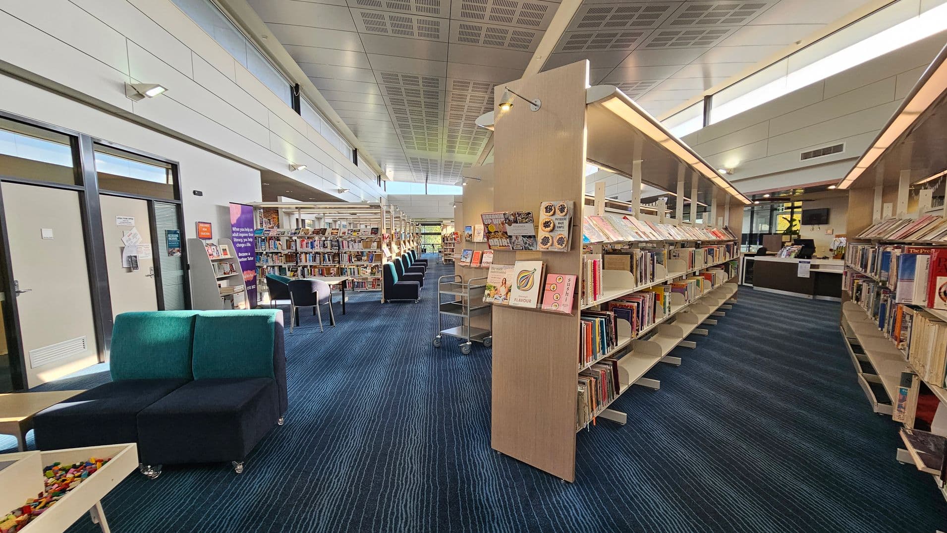 A modern library interior with a blue-striped carpet. On the left, there are glass doors and a seating area with teal and black chairs. In the centre, bookshelves display various books and magazines, with a trolley nearby. On the right, more bookshelves lead to a reception desk area. The ceiling is high with a grid pattern and skylights, allowing natural light to illuminate the space.