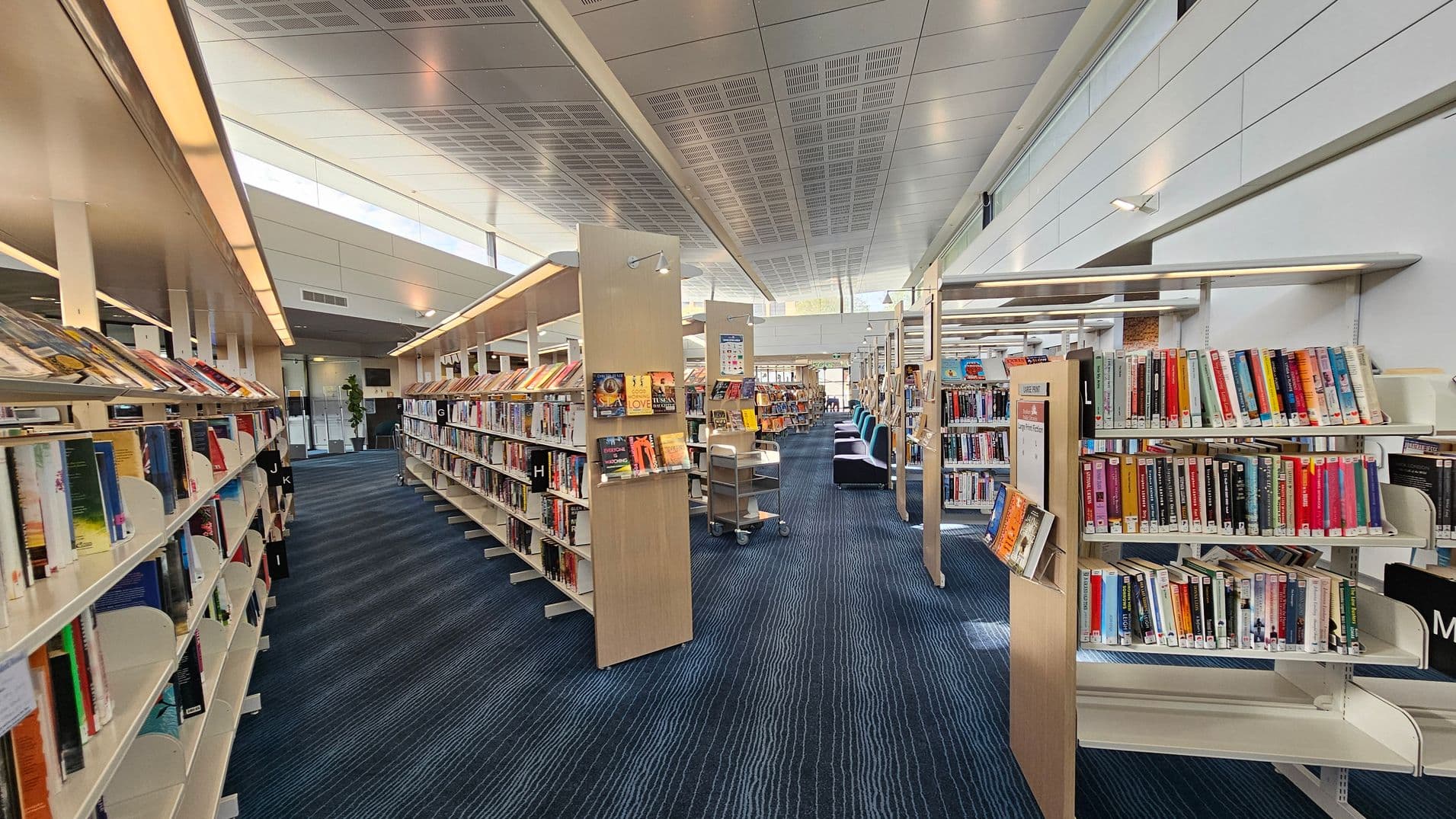 A modern library interior with rows of bookshelves on both sides. The shelves are filled with books, organised alphabetically with visible letters like H, I, J, K, L, and M. The floor is covered with a blue carpet featuring subtle striped patterns. In the background, there are more shelves and a few comfortable chairs. The ceiling is high with a grid pattern and large windows allowing natural light to illuminate the space.
