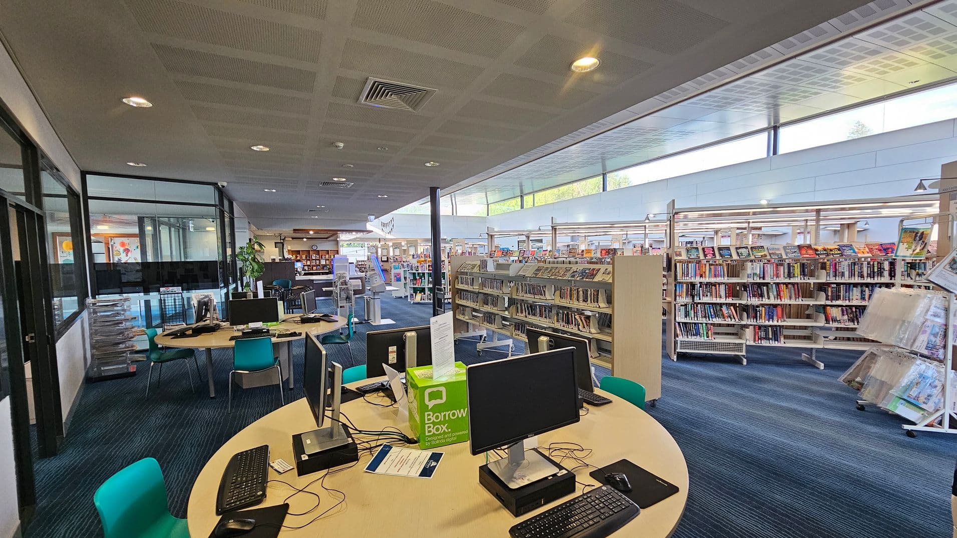 A library interior with a modern design. On the left, there are round tables with computers and teal chairs. The floor is carpeted in blue with a striped pattern. In the middle, there are bookshelves filled with books, and a "Borrow Box" is visible on a table. The right side features more bookshelves and a display rack with magazines. The ceiling is fitted with recessed lighting and air vents, and large windows allow natural light to illuminate the space.