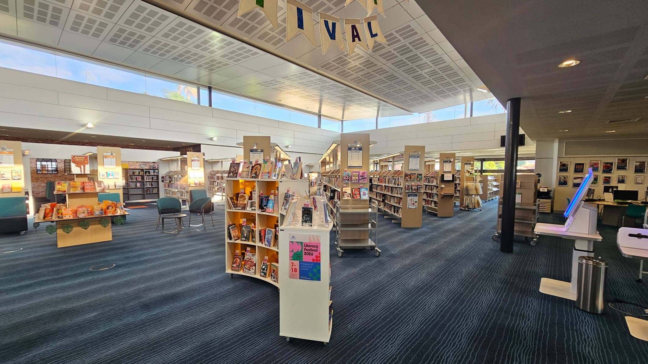A spacious library interior with a high ceiling featuring grid-like patterns. In the centre, there are curved bookshelves filled with books, and a sign for a "Literacy Festival 2026". To the left, more bookshelves and a table with orange-themed books are visible. On the right, a digital kiosk with a glowing screen stands near a black pillar. The floor is carpeted in blue with subtle patterns, and large windows allow natural light to illuminate the space.