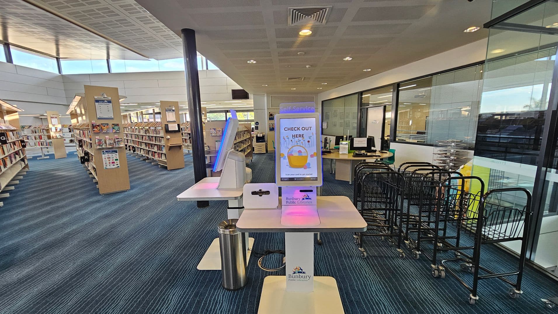 A library interior with a self-service checkout station in the centre, featuring a screen displaying "CHECK OUT HERE." To the right, there are several book trolleys and a glass wall with a view of an office area. On the left, rows of bookshelves are filled with books. The floor is covered with blue carpet, and the ceiling has a grid pattern with recessed lighting.