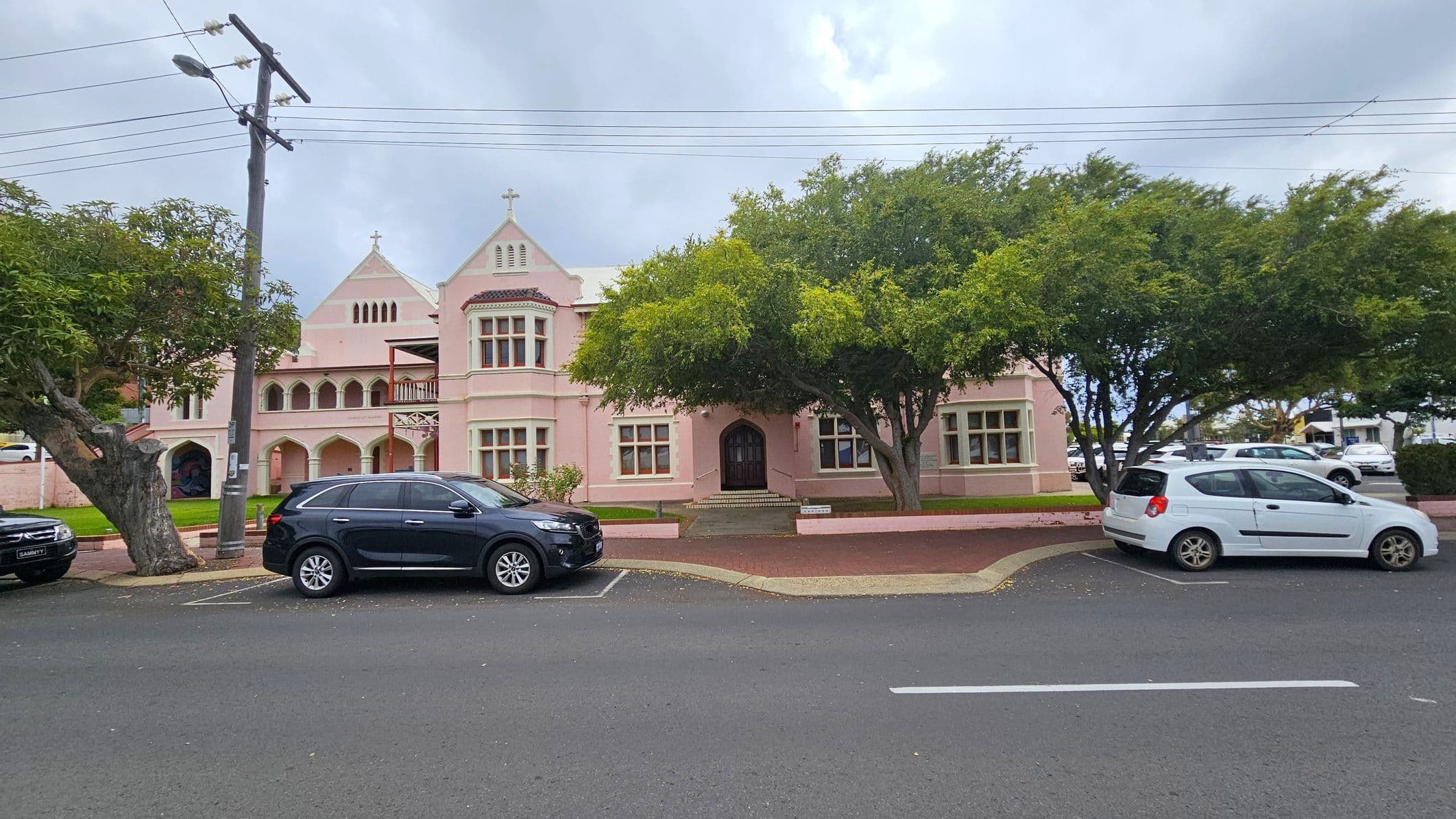 A pink, two-storey building with Gothic architectural elements, including arched windows and a cross on the roof. In front, there are two large trees partially obscuring the facade. A black SUV and a white car are parked on the street, which has a marked parking area. A power pole with wires is on the left. The pavement is red brick, and the road is asphalt.