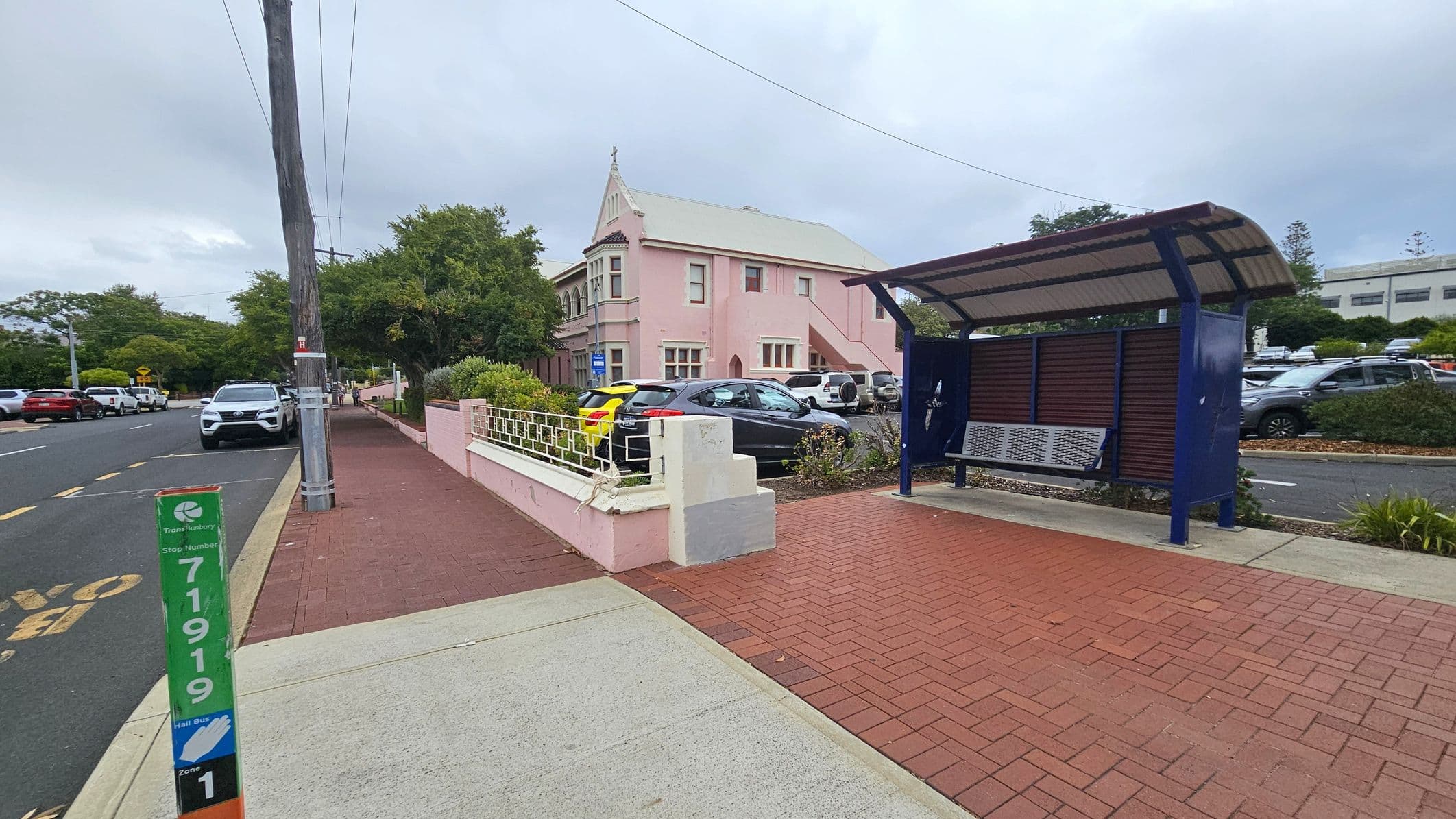 A street scene with a pink building on the left, featuring a gabled roof and multiple windows. In front of the building, several cars are parked along the street. A bus stop with a blue frame and a metal bench is on the right, set on a red brick pavement. A green and orange bus stop sign is visible in the foreground. The street is lined with trees and a power pole, and the pavement is a mix of red bricks and concrete.