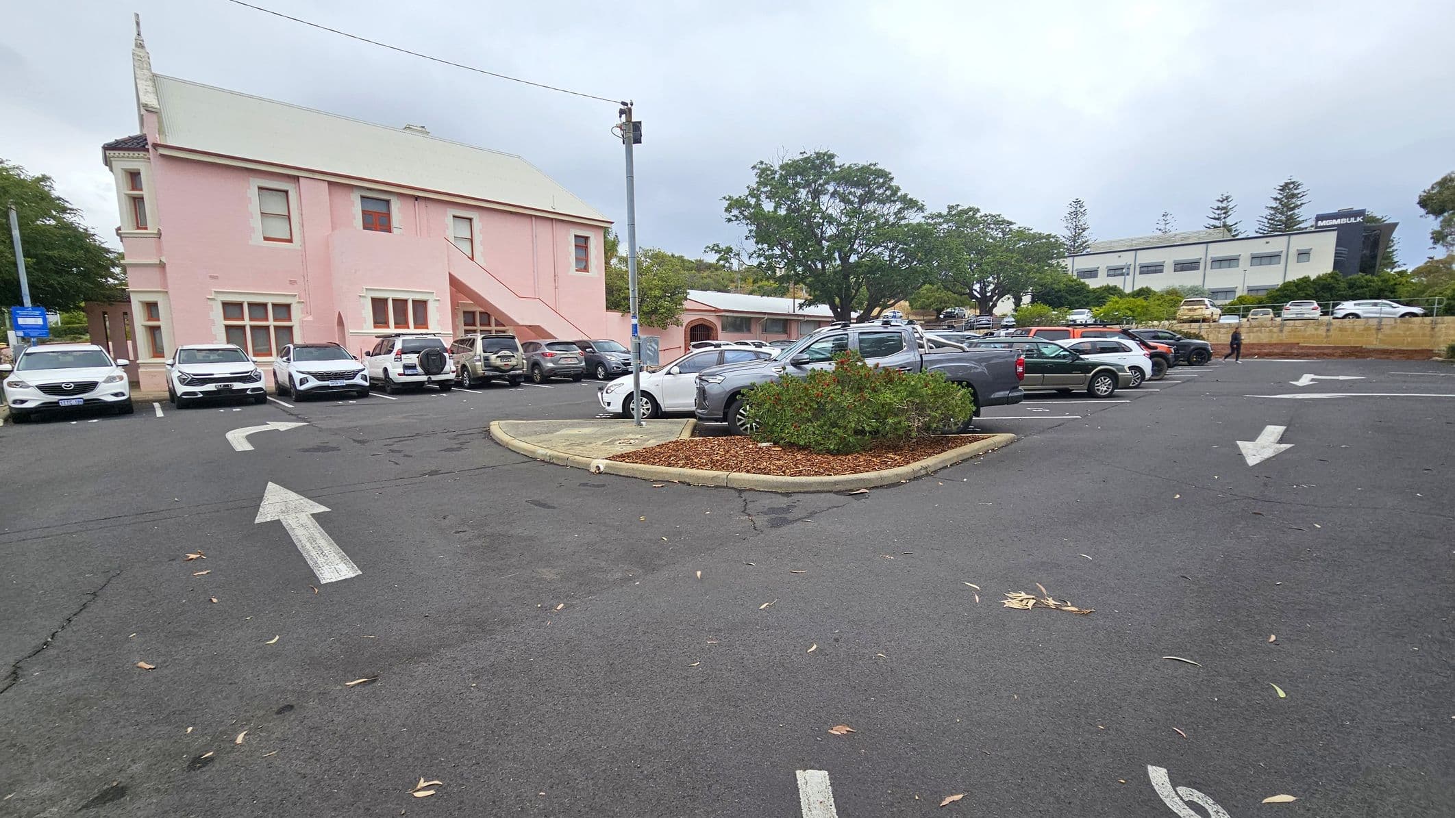 A car park with several vehicles is visible. On the left, there's a pink building with a sloped roof and external staircase. In the centre, a small landscaped area with shrubs is surrounded by parked cars. On the right, more cars are parked, and a person is walking. In the background, a white building with the word "MSMBULK" is partially visible, along with trees and a low wall. The asphalt surface has directional arrows painted on it.