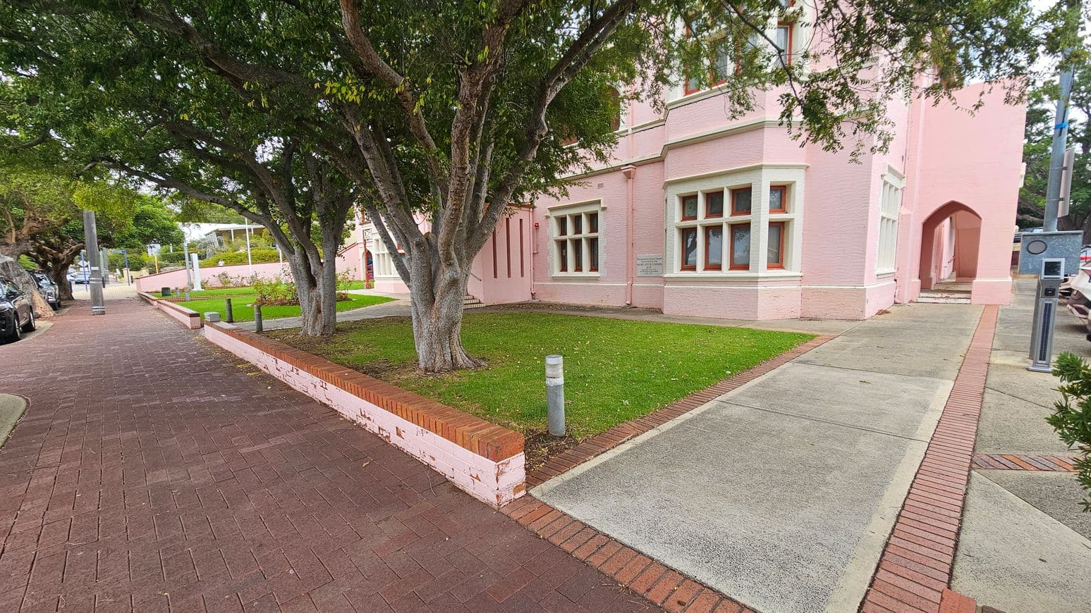A pink brick building with large windows is on the right, featuring an arched entrance. In the foreground, a green lawn with a tree is bordered by a low brick wall. The left side has a paved footpath lined with trees and parked cars. A parking meter is visible near the building. The pavement is a mix of red and grey bricks.