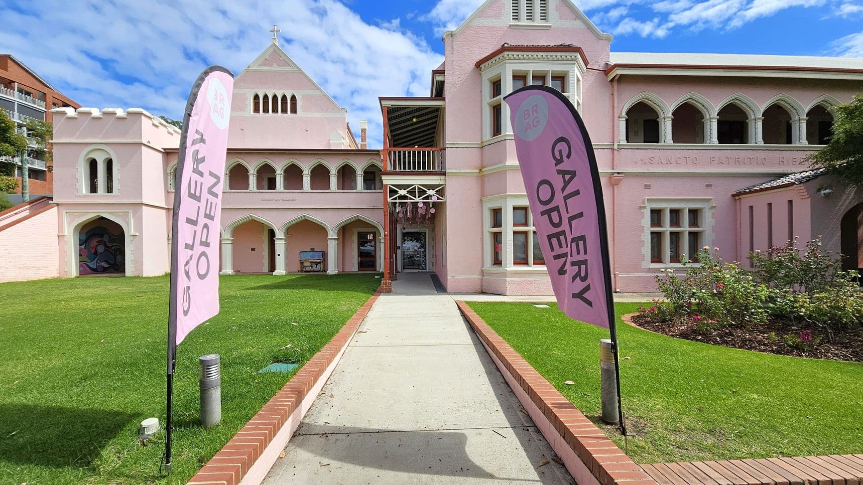 A pink building with Gothic-style architecture, featuring arched windows and a cross on the roof. Two vertical banners on either side of a concrete path read "GALLERY OPEN". The path is bordered by red brick edging and leads to the entrance. The left side of the building has a mural under an archway. The right side has a garden with shrubs and flowers. The grass is neatly trimmed, and the sky is partly cloudy.