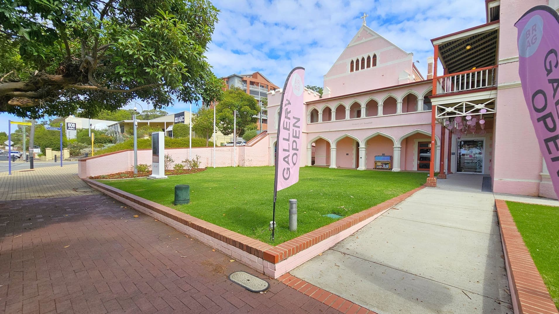 A pink building with arched windows and a cross on top is on the right, with a sign reading "GALLERY OPEN" on a flag. A paved path leads to the entrance, bordered by a green lawn and low brick walls. On the left, a tree overhangs a brick pavement. In the background, there are signs for "ABC" and other buildings, with a few cars visible.
