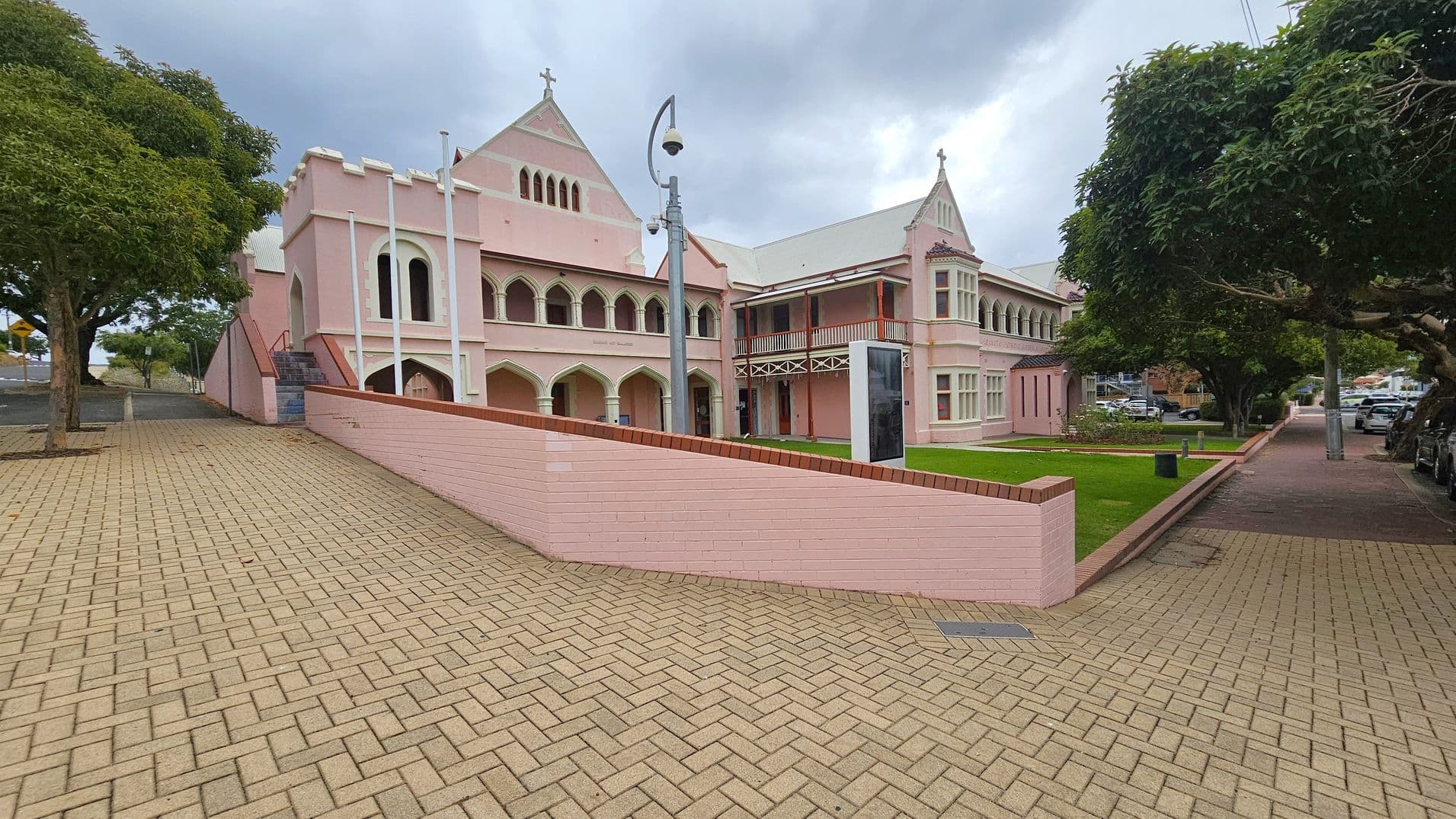 A pink building with Gothic architectural elements, including arched windows and a cross on the roof, is situated in the centre. The foreground features a paved area with a herringbone pattern. On the left, a tree-lined path leads to the building's entrance, while on the right, a grassy area with more trees and parked cars lines the street. A modern streetlamp stands near the building.