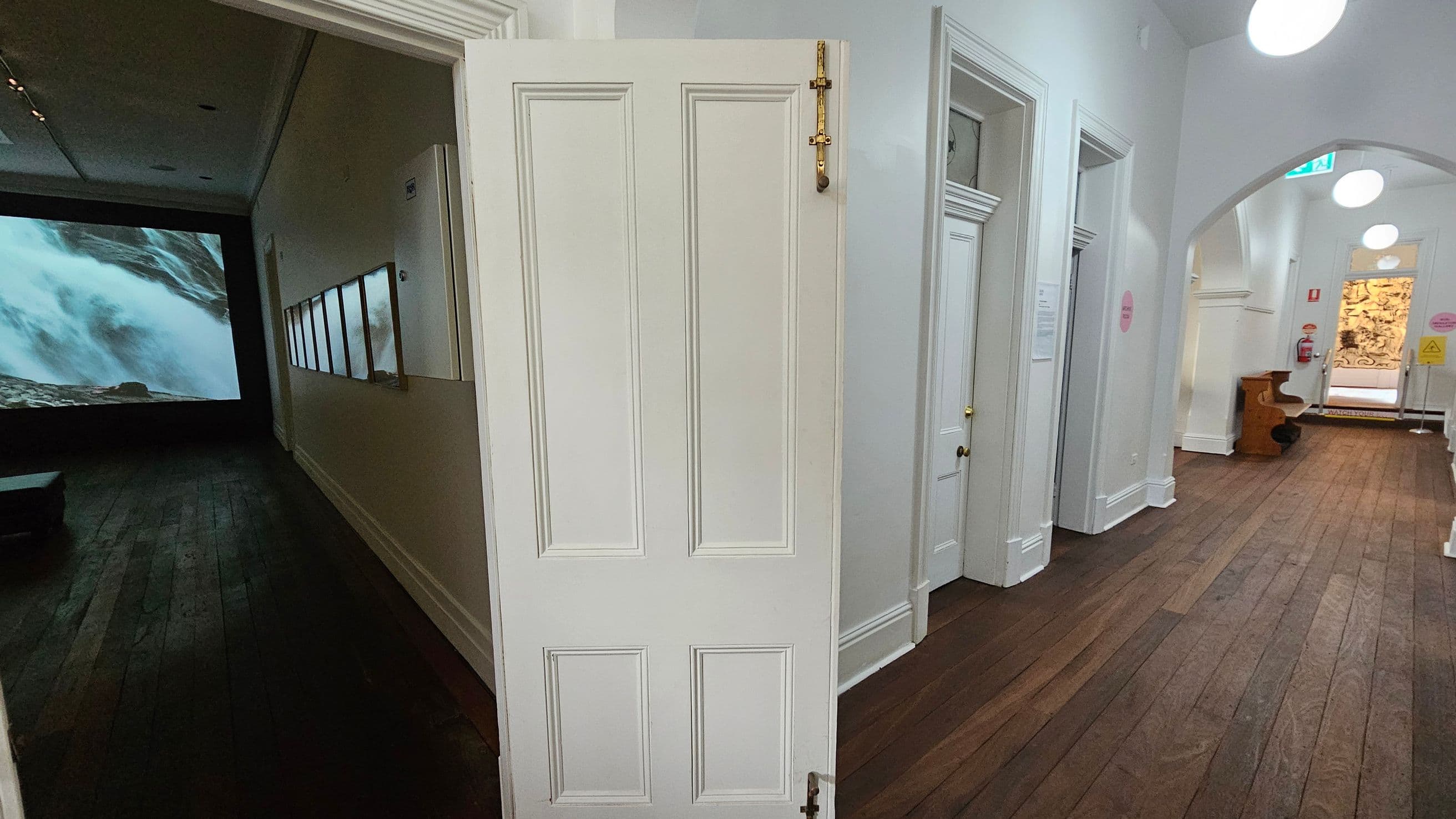 A hallway with wooden flooring and white walls. On the left, an open door reveals a dark room with a large screen displaying a waterfall. The hallway extends to the right, featuring several closed white doors with brass handles. The ceiling has round, white lights. At the end of the hallway, there's a bench and a fire extinguisher on the wall. An exit sign is visible above an arched doorway leading to another area with a decorative panel.