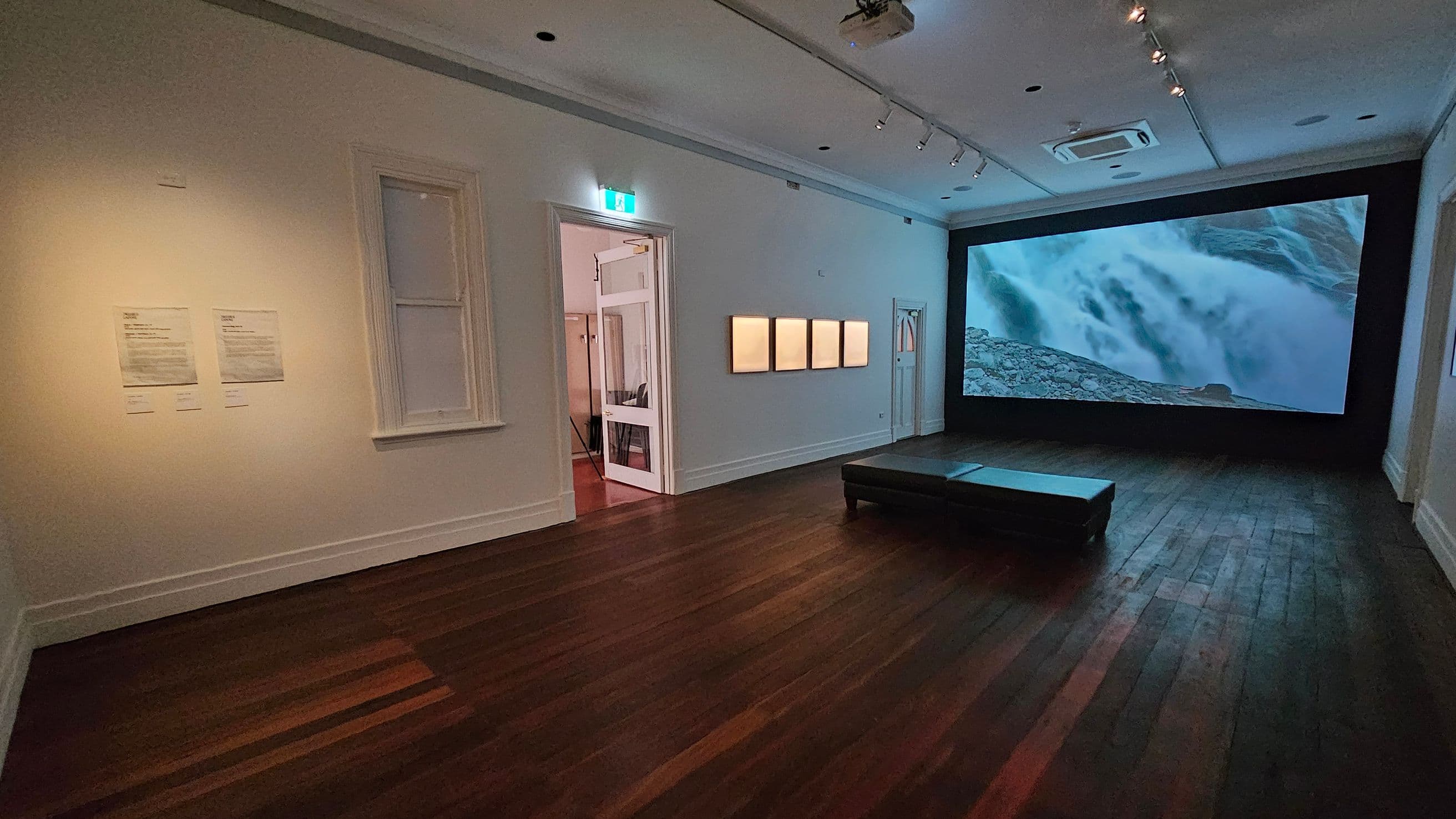 A gallery room with wooden flooring and a white ceiling featuring recessed lighting. On the right, a large screen displays an image of a waterfall. In the centre, a black bench is positioned on the floor. The left wall has two framed documents and a window. An open doorway leads to another room, with a lit exit sign above. Four framed artworks are mounted on the wall near the doorway.
