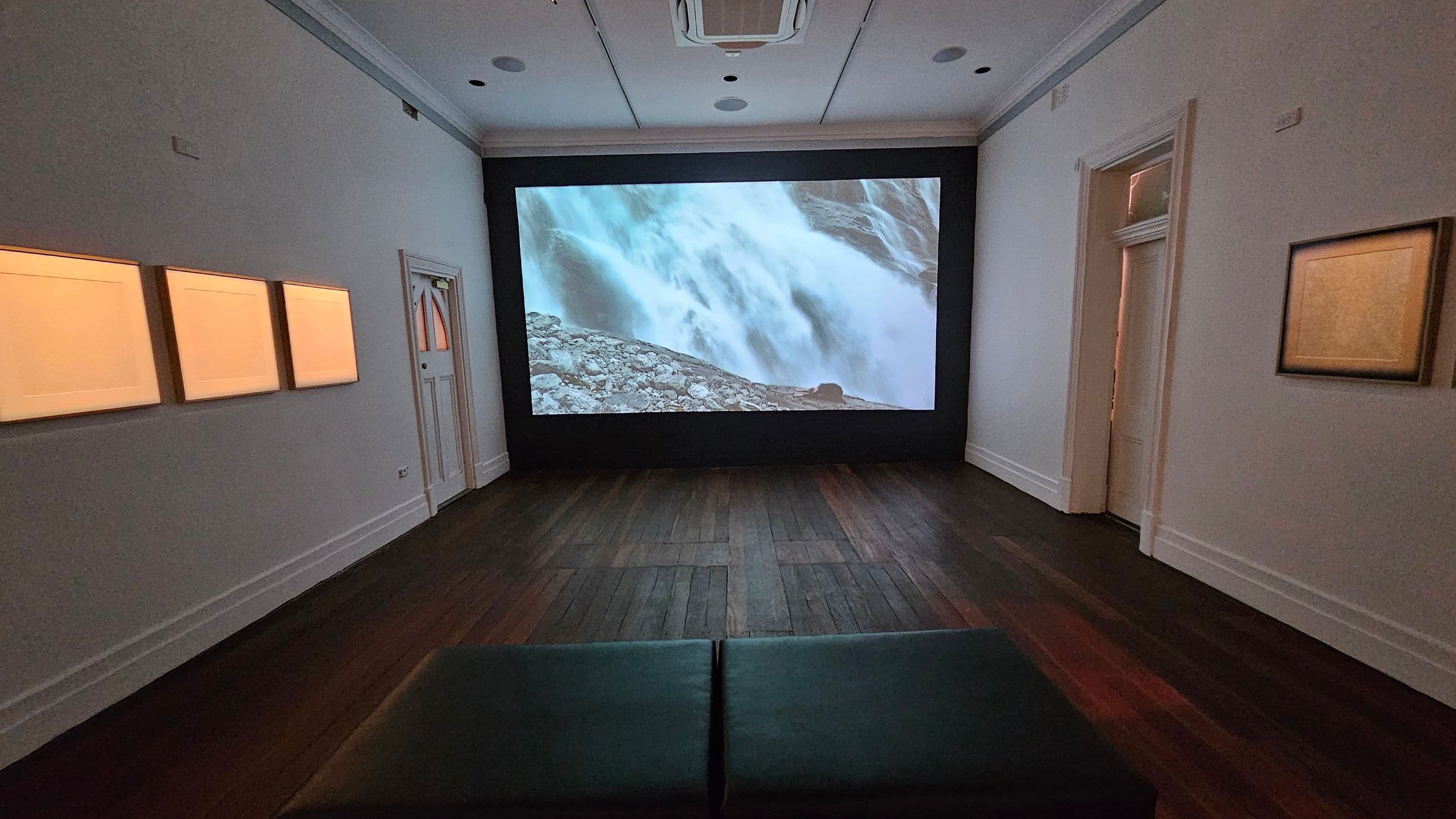 A room with dark wooden flooring and a white ceiling featuring recessed lights and an air conditioning unit. In the centre, a large screen displays an image of a waterfall. On the left wall, three illuminated rectangular panels are mounted. On the right wall, a single framed piece is visible. A doorway is present on the right side. In the foreground, there is a dark bench facing the screen.