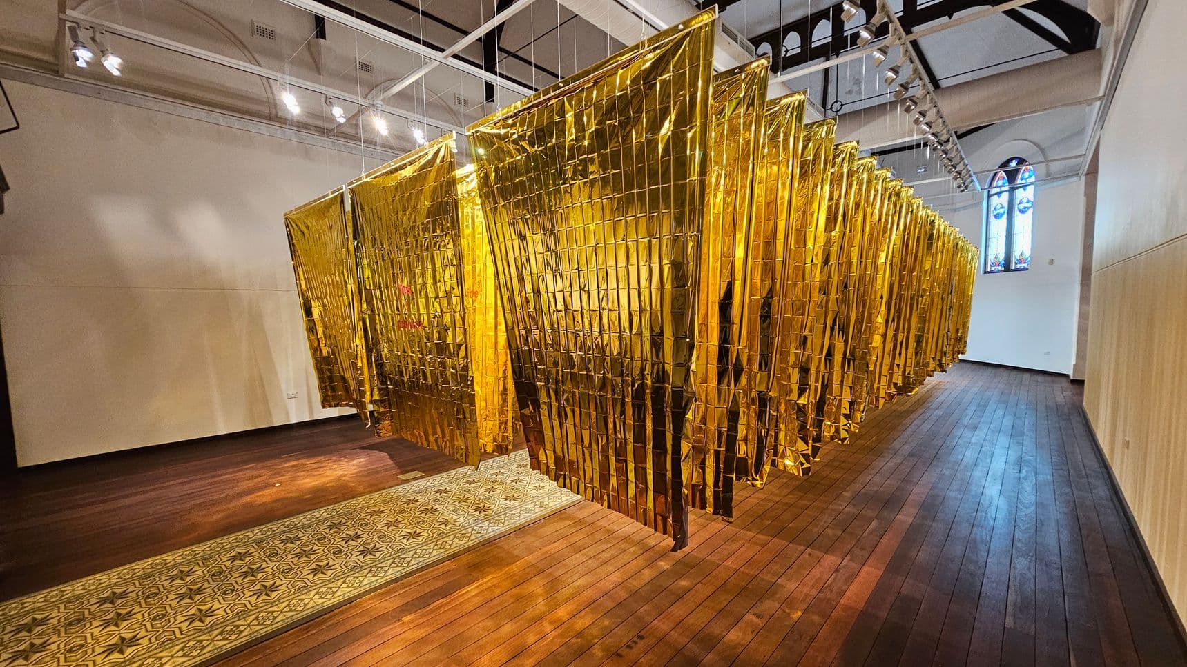 A room with a wooden floor and a high ceiling featuring exposed beams and spotlights. In the centre, a series of golden, reflective sheets hang in a zigzag pattern from the ceiling. On the left, a plain white wall, while on the right, a stained glass window adds colour to the space. A patterned carpet runs along the floor beneath the golden sheets.