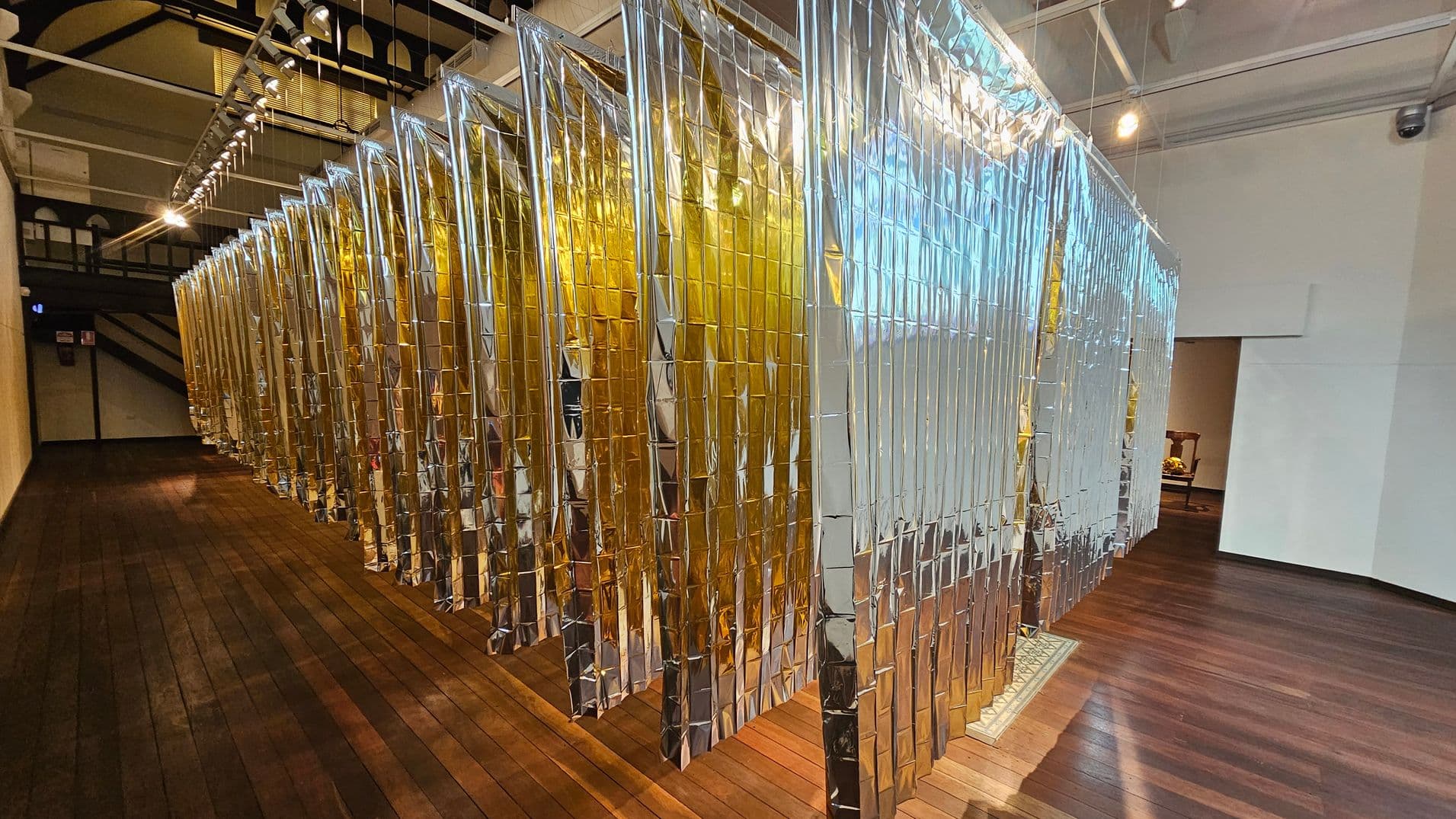 A room with a wooden floor and a high ceiling featuring a series of metallic, reflective panels hanging from above. The panels are arranged in a row, alternating between silver and gold colours. On the left, the panels are illuminated by overhead lights, casting reflections on the floor. The right side shows a white wall with an open doorway leading to another room. The ceiling has exposed beams and track lighting.
