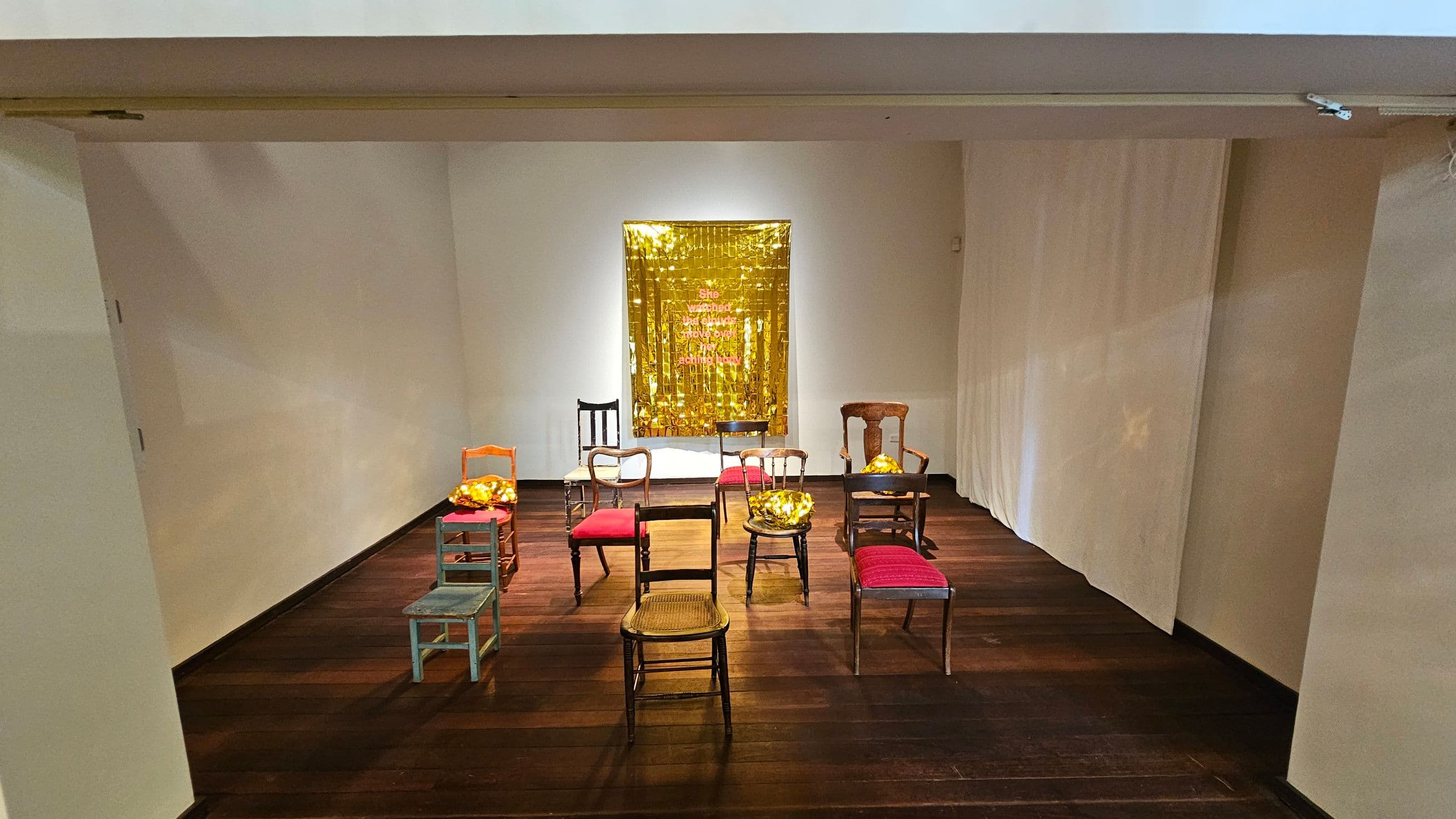 A room with a wooden floor and a white ceiling. In the centre, there are several chairs arranged in a semi-circle, each with a cushion. The chairs vary in style and colour, with some having red seats. On the back wall, a gold, reflective curtain hangs with text on it. The walls are plain and light-coloured, and the space is well-lit.