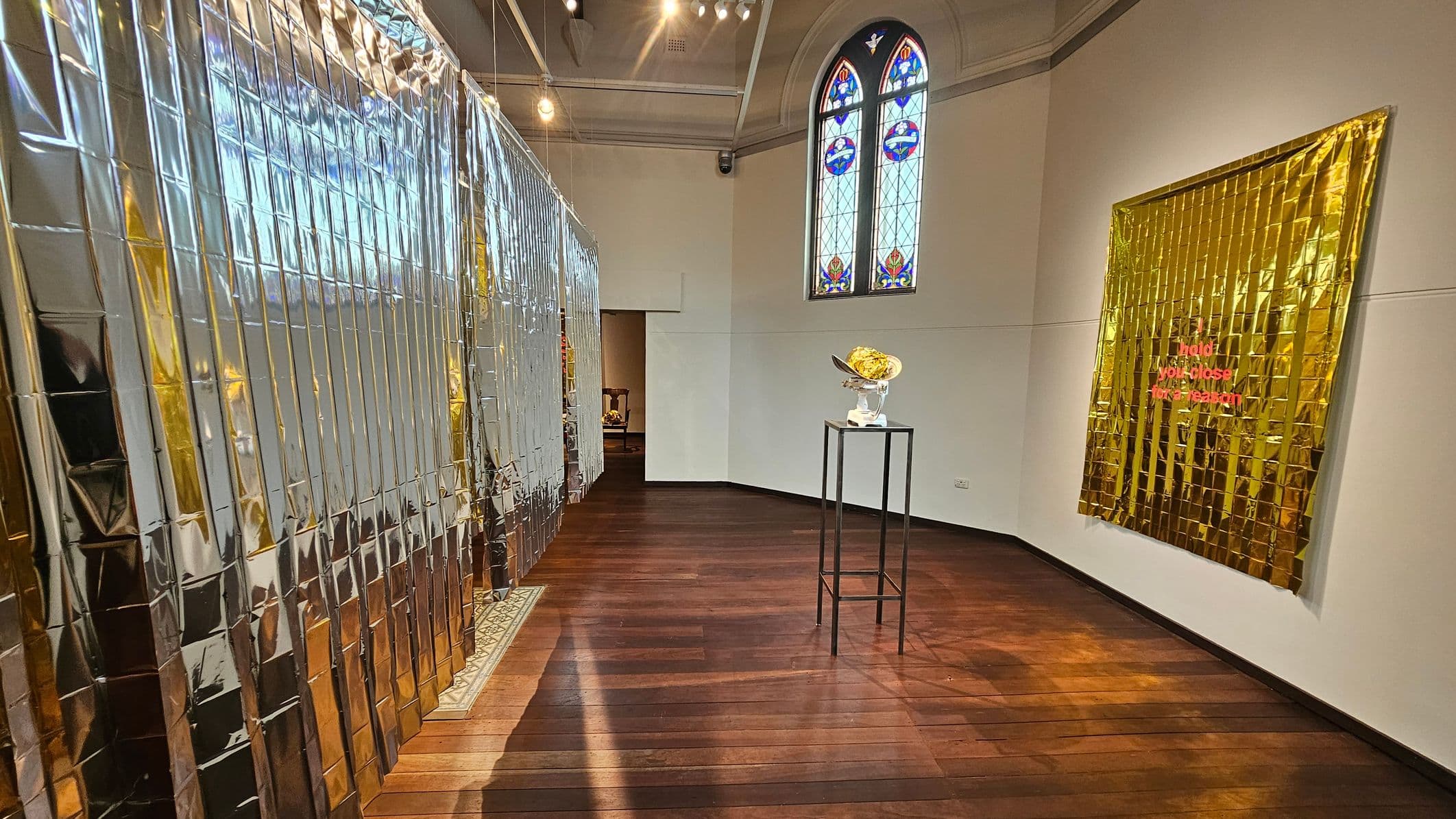A room with dark wooden flooring and a high ceiling. On the left, a wall is covered with reflective metallic sheets. In the centre, a tall black stand holds a sculptural piece. On the right, a golden metallic sheet with red text is mounted on the wall. In the background, a stained glass window with colourful patterns is visible. The lighting is warm, creating reflections on the floor.