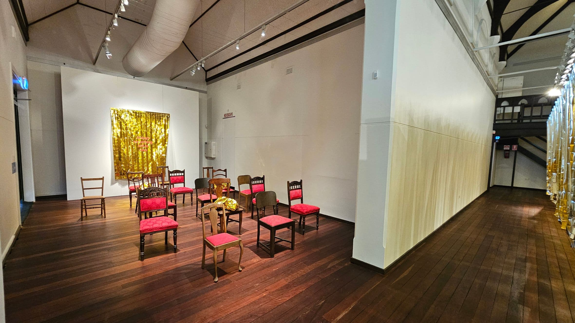 A room with wooden flooring and a high ceiling featuring exposed beams and a large duct. On the left, a group of chairs with red cushions is arranged in a semi-circle. Behind them, a gold, reflective wall hanging is displayed. On the right, a long corridor with a light-coloured wall and a series of vertical, metallic strips hanging from the ceiling. The lighting is provided by spotlights along the ceiling.