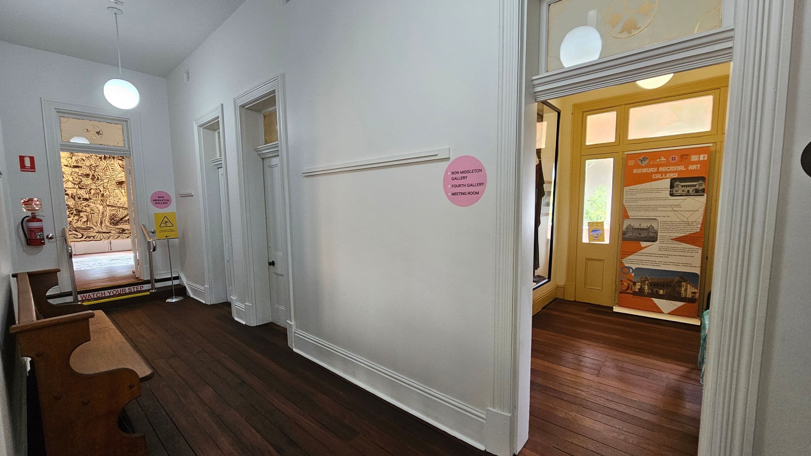 A hallway with wooden flooring and white walls. On the left, there's a wooden bench and a fire extinguisher mounted on the wall. A door with a "Watch Your Step" sign leads to a room with a mural. On the right, a doorway opens to a space with a large informational banner about the Sunbury Regional Art Gallery. The ceiling features hanging round lights. A pink sign on the wall indicates directions to various galleries and a meeting room.