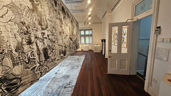 A room with a large, detailed black and white mural covering the left wall. The floor is dark wood, and a long, light-coloured rug runs along it. On the right, an open door leads to another room with framed artworks on the wall. The ceiling is white with exposed beams and track lighting. At the far end, a window with multiple panes is flanked by two small pedestals. A dark fireplace is visible on the right wall.