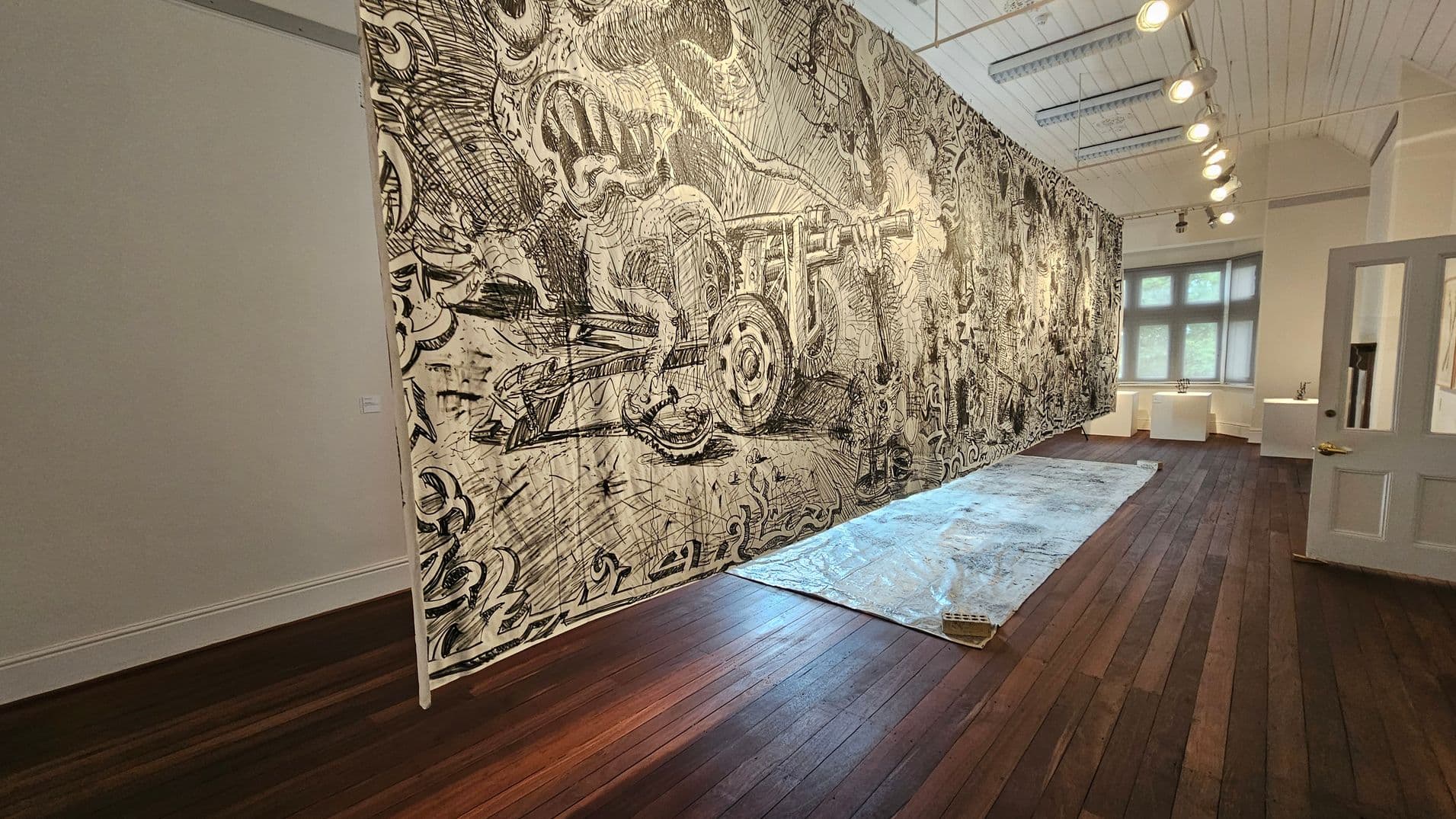 A room with dark wooden flooring and a high, white panelled ceiling. On the right, a large, detailed black and white mural covers the wall, depicting intricate, abstract designs. A plastic sheet is spread on the floor in front of the mural. In the background, a window with multiple panes allows natural light into the room. To the right, a partially open door leads to another space. Ceiling lights illuminate the mural.