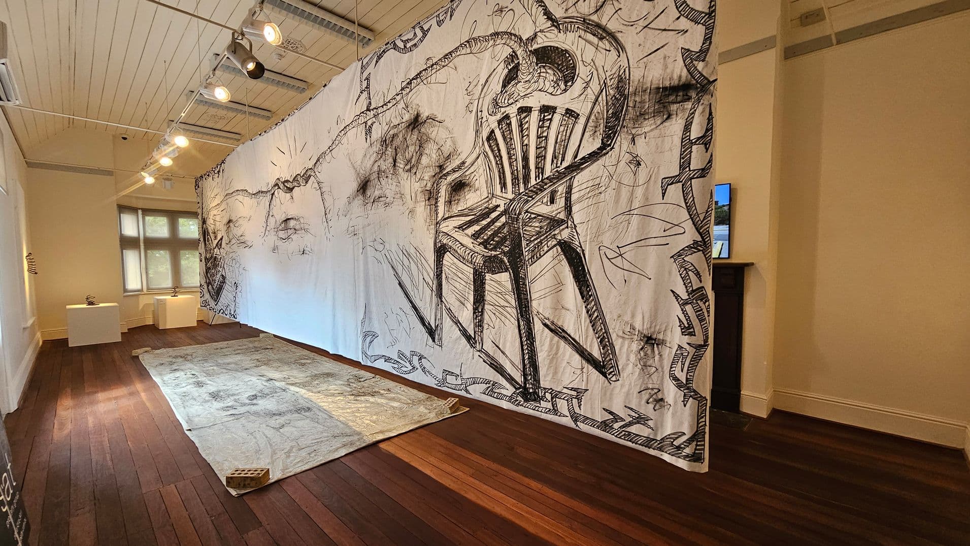 A room with wooden flooring and a white panelled ceiling. A large, intricately drawn artwork featuring a chair and abstract designs hangs prominently in the centre. On the left, two small sculptures rest on white pedestals near a window. A textured rug lies on the floor with a brick placed on one corner. Track lighting illuminates the space, and a small screen is visible on the right wall.