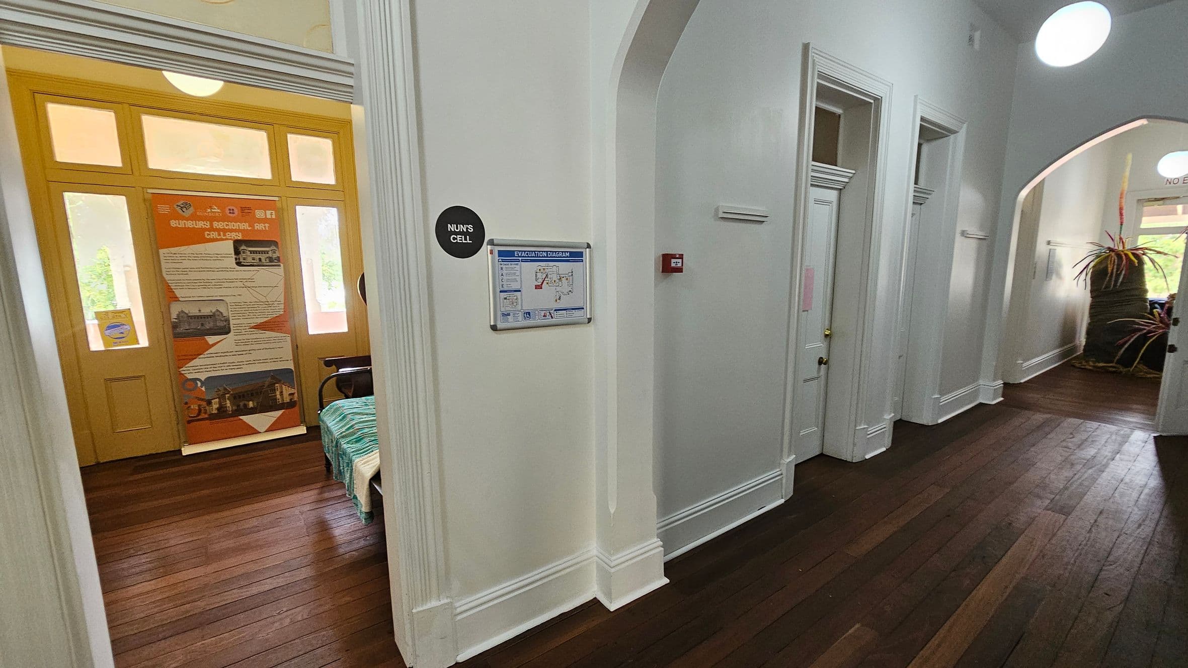 A hallway with wooden flooring and white walls. On the left, a room labelled "Nun's Cell" has a yellow door with a poster about the Sunbury Regional Art Gallery. In the centre, an evacuation diagram is mounted on the wall. On the right, there are several closed white doors and a large plant in a pot near an arched opening. Circular lights are visible on the ceiling.