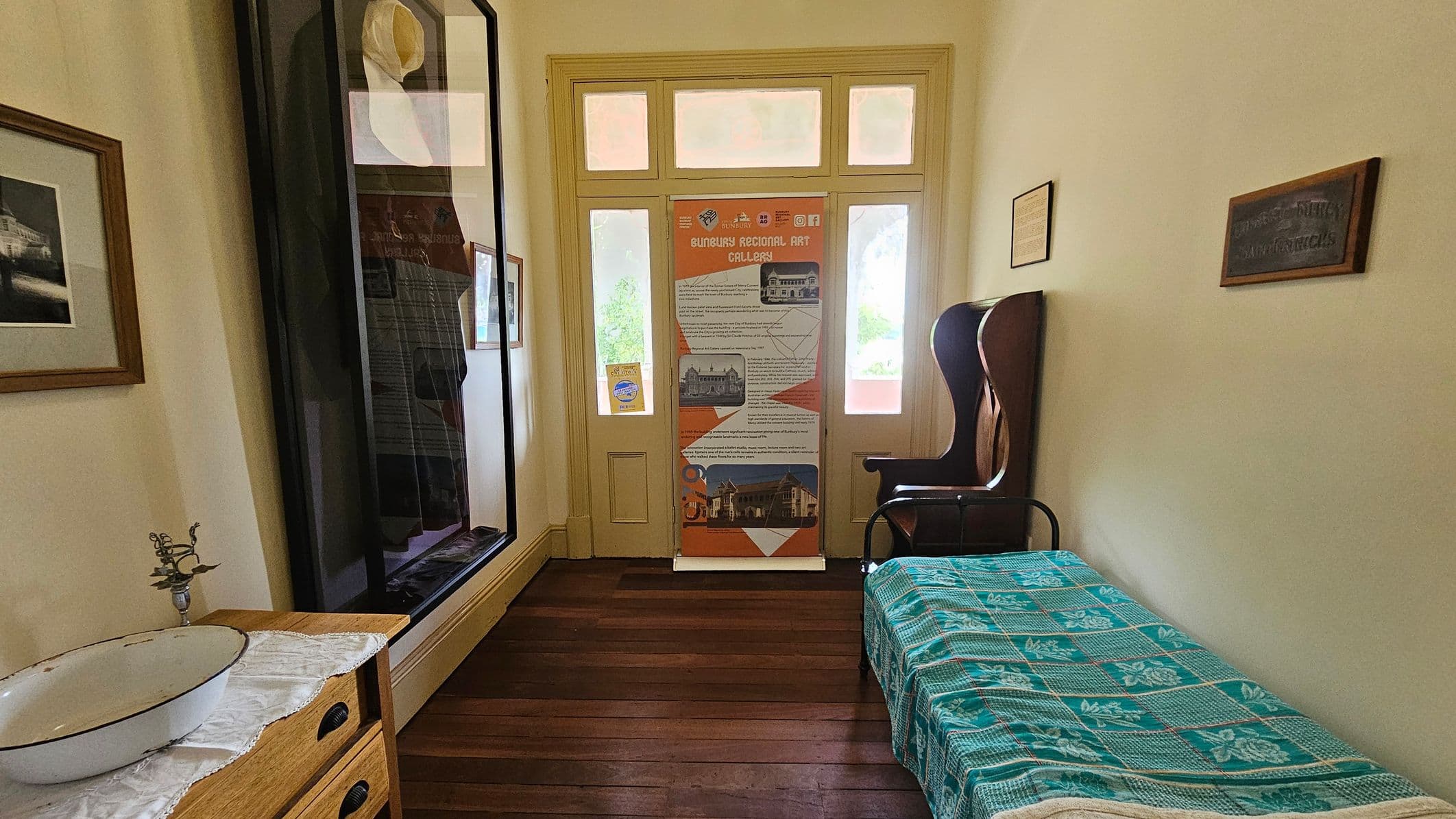 A small room with wooden flooring and cream-coloured walls. On the left, there's a wooden dresser with a white basin and a decorative metal stand. Above it, a framed picture hangs on the wall. A large framed display case is next to the dresser. In the centre, a door with glass panels and a colourful informational banner. On the right, a single bed with a blue patterned cover and a wooden chair. A small framed sign is mounted on the wall above the bed.