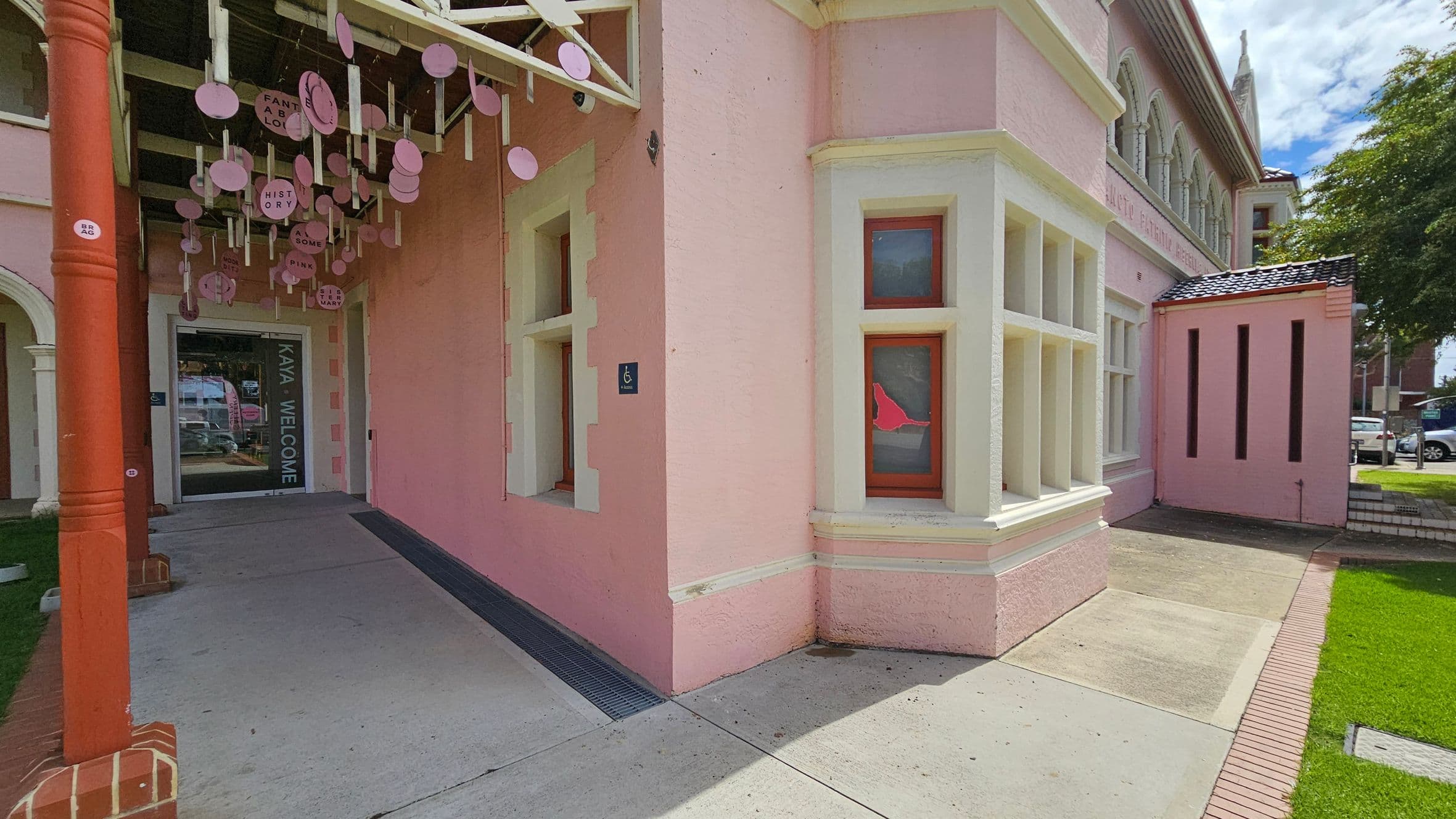 A pink building with white trim features a corner entrance. On the left, a covered walkway with hanging circular decorations leads to a glass door with "KAYA WELCOME" written on it. The right side shows a series of windows with red frames. The ground is a mix of concrete and grass, with a small brick border. A tree and parked cars are visible in the background.