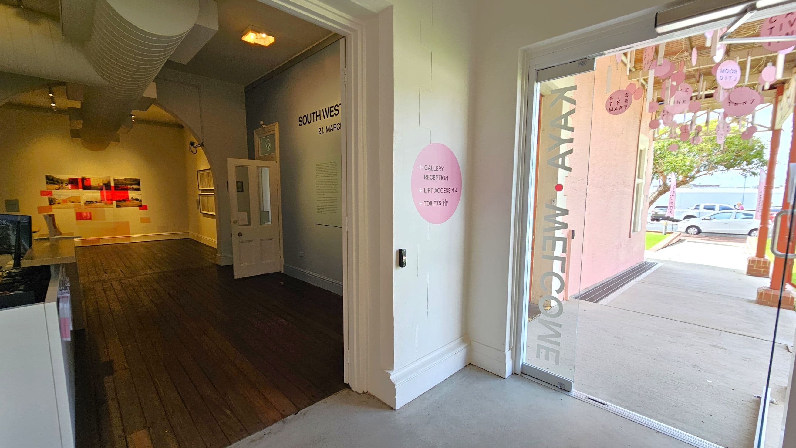 A gallery entrance with a glass door on the right, displaying the words "KAYA WELCOME". Outside, there are pink hanging decorations and parked cars visible. Inside, on the left, is a room with wooden flooring and artwork on the walls. A sign on the wall indicates directions to the gallery reception, lift access, and toilets. The ceiling features exposed ductwork and lighting fixtures.