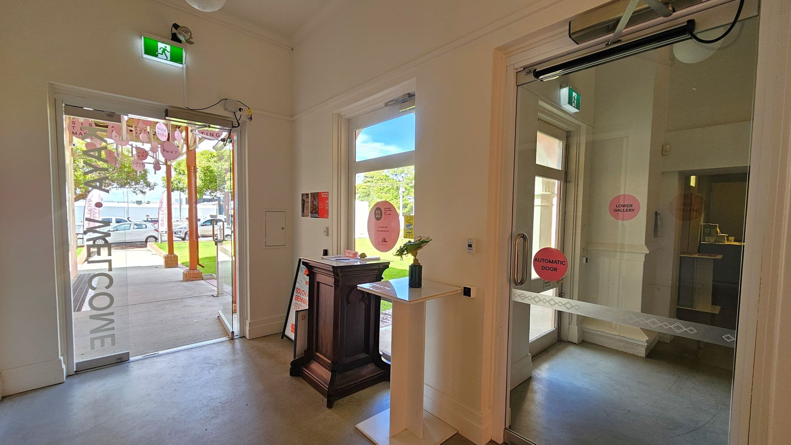 A room with two glass doors. On the left, an open door leads outside, showing a grassy area and parked cars. Above it, a green exit sign is visible. On the right, a closed automatic door with signs reading "Automatic Door" and "Lower Gallery". Between the doors, a small table with a vase and a wooden stand with brochures. The floor is light-coloured, and the walls are white.
