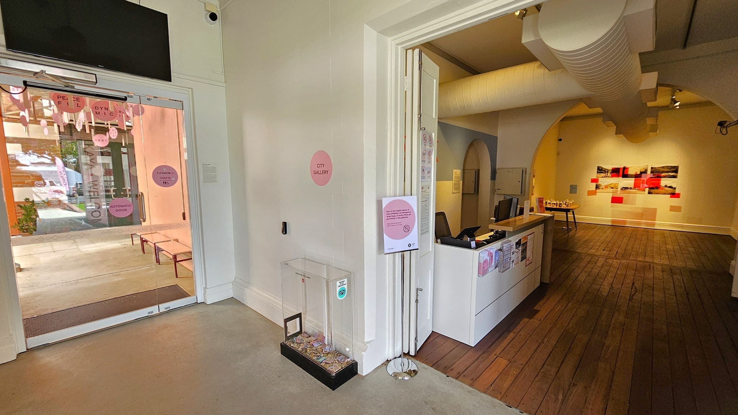 A gallery entrance with a glass door on the left, displaying signs and a view of the outside with benches and greenery. Inside, a donation box is placed near the door. On the right, a reception desk with brochures and a computer is situated on a wooden floor. The room has a large duct on the ceiling and an archway leading to a wall with colourful artwork. The walls are light-coloured, and there is a sign on a stand near the entrance.